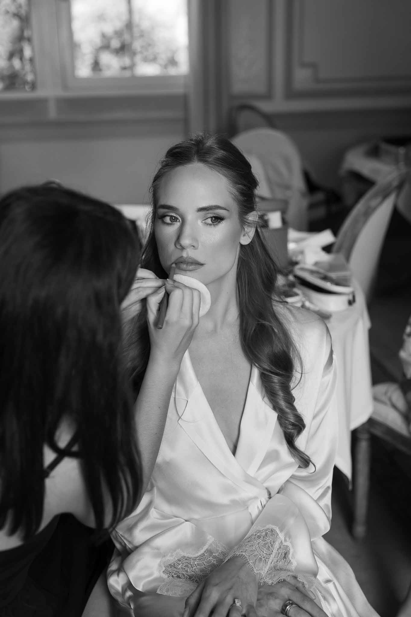 Black-and-white portrait of bride seated indoors while makeup artist applies powder with a sponge puff
