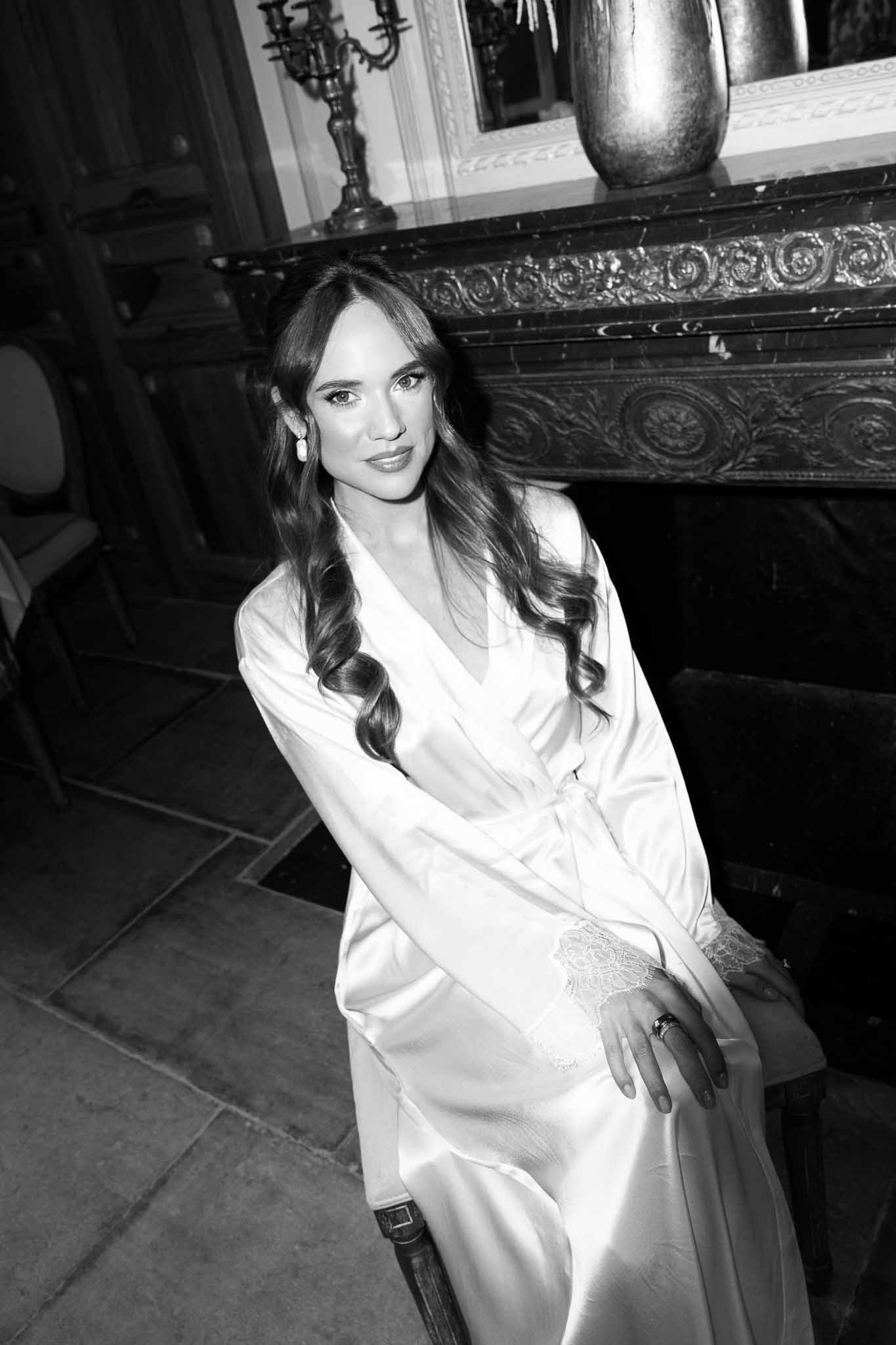 Black and white portrait of bride in satin robe seated before carved marble fireplace