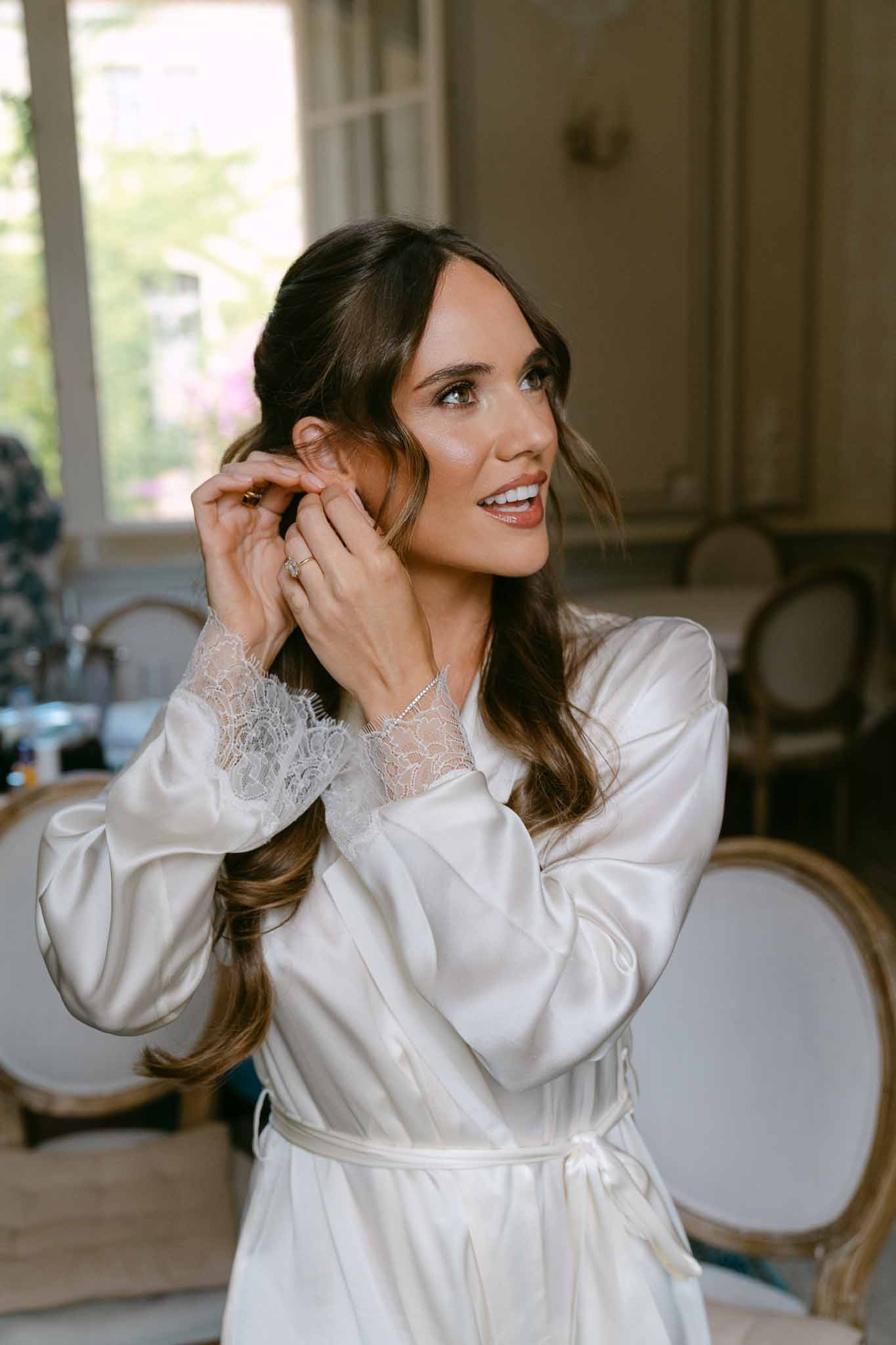 Bride smiling while putting on earring in satin robe inside ornate French chateau room during getting ready