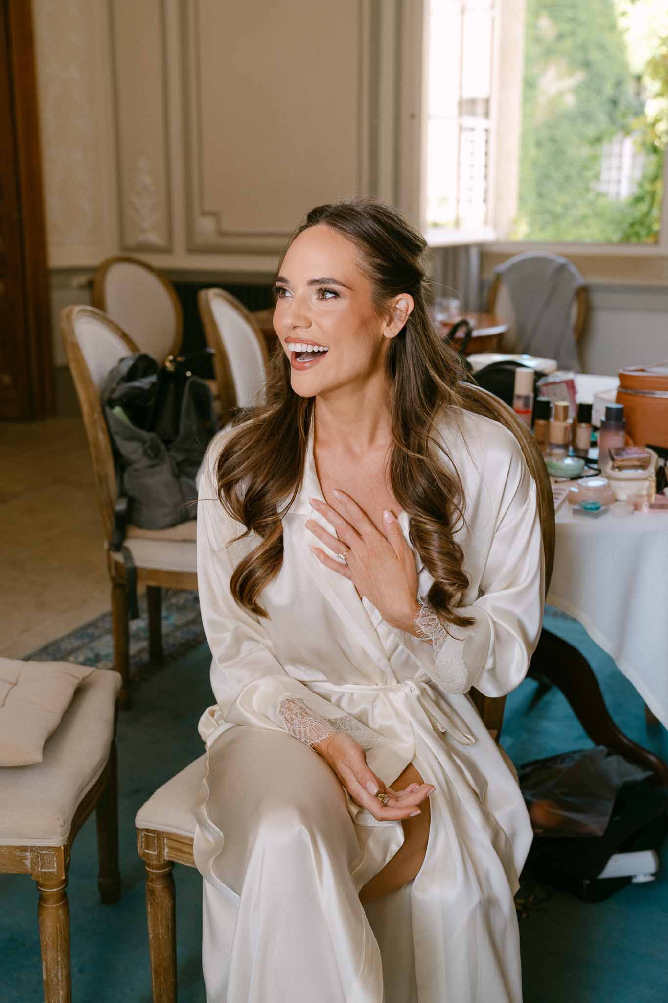 Bride in cream satin robe with voluminous waves laughing on French chair in panelled chateau room