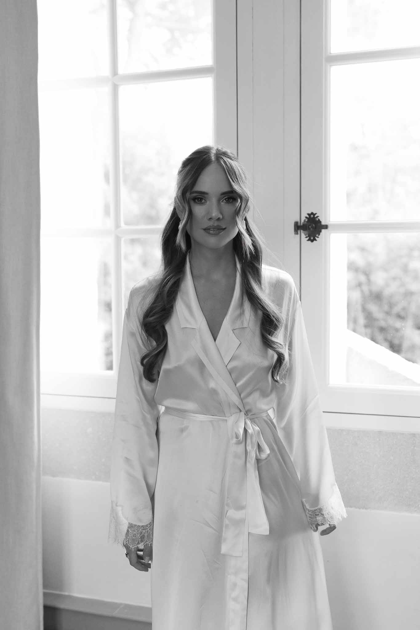 Black and white bride in satin robe with lace cuffs standing before French windows with bathtub behind