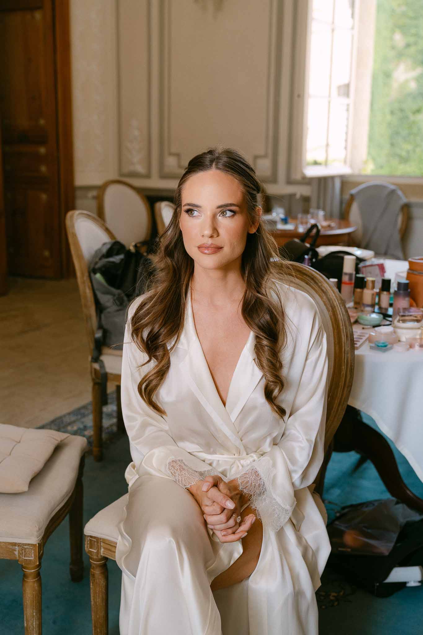 Bride in ivory satin robe during hair and makeup preparations in chateau room