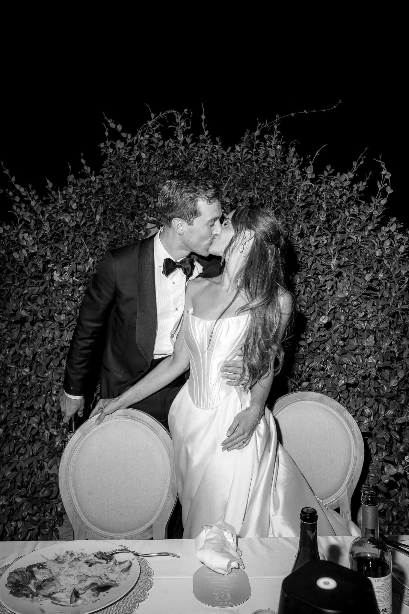 Black and white flash portrait of couple kissing at reception table with hedge backdrop at night