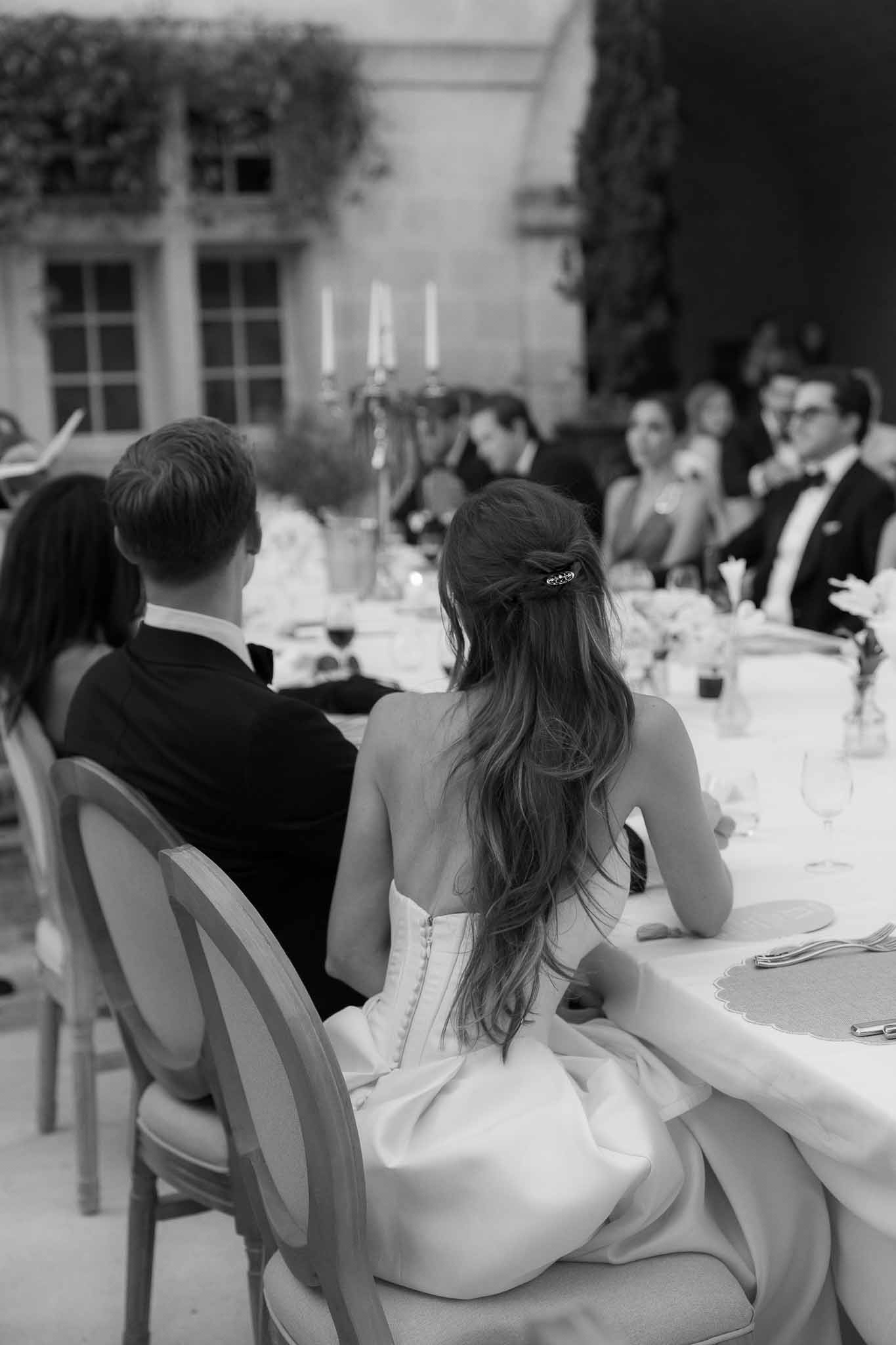 Black and white photo of bride and groom seated at reception table with candelabra and guests in courtyard