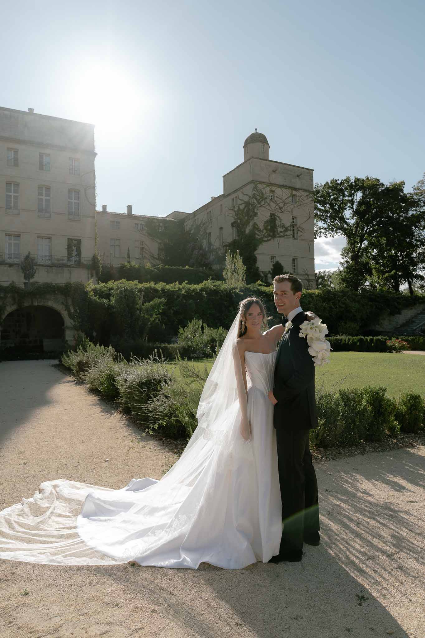 Bride in strapless gown with cathedral veil and groom in navy suit in formal chateau garden with domed tower