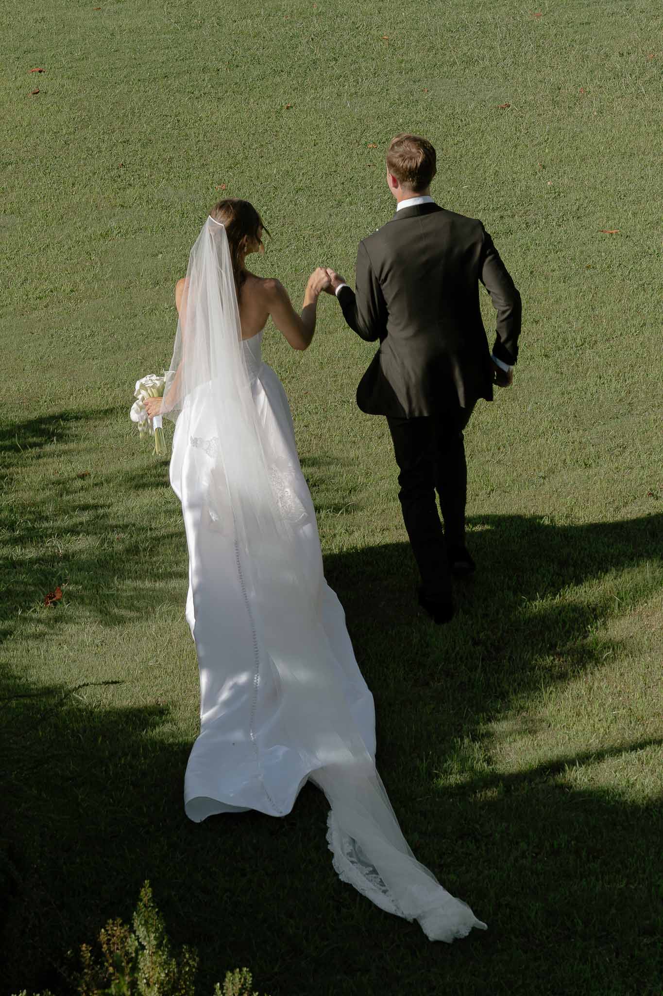 Elevated view of couple walking across lawn from behind with cathedral train and veil spread across grass