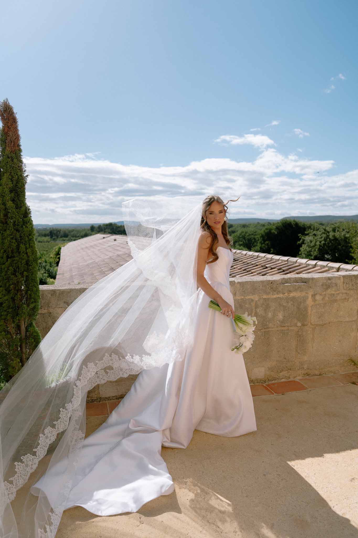 Bride on stone terrace with wind-blown cathedral lace veil holding white calla lily bouquet and countryside beyond