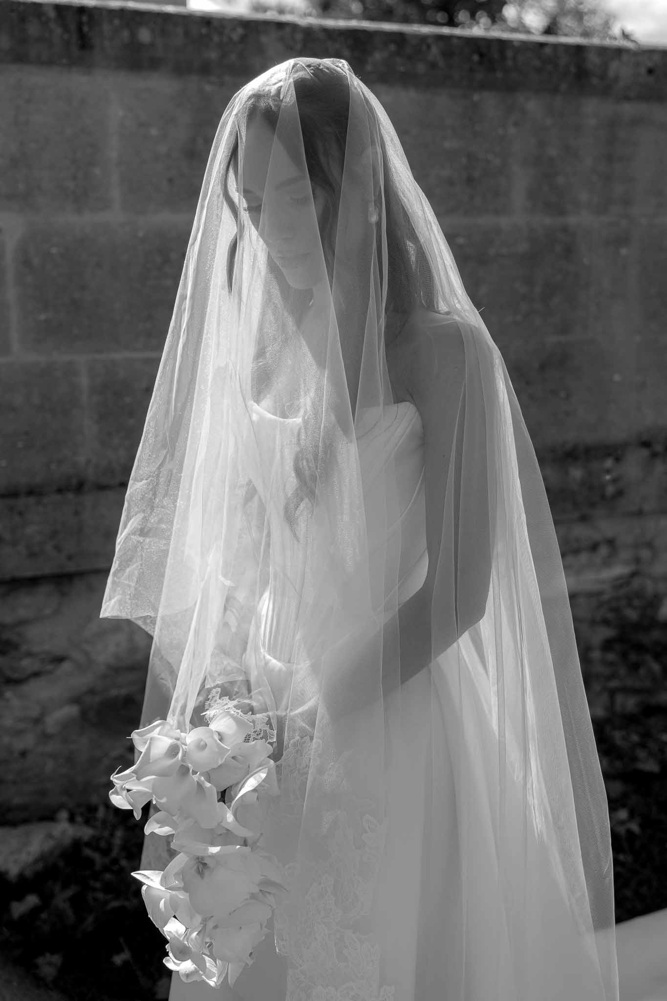 Black and white portrait of bride with sheer veil over face holding cascading calla lily bouquet by stone wall