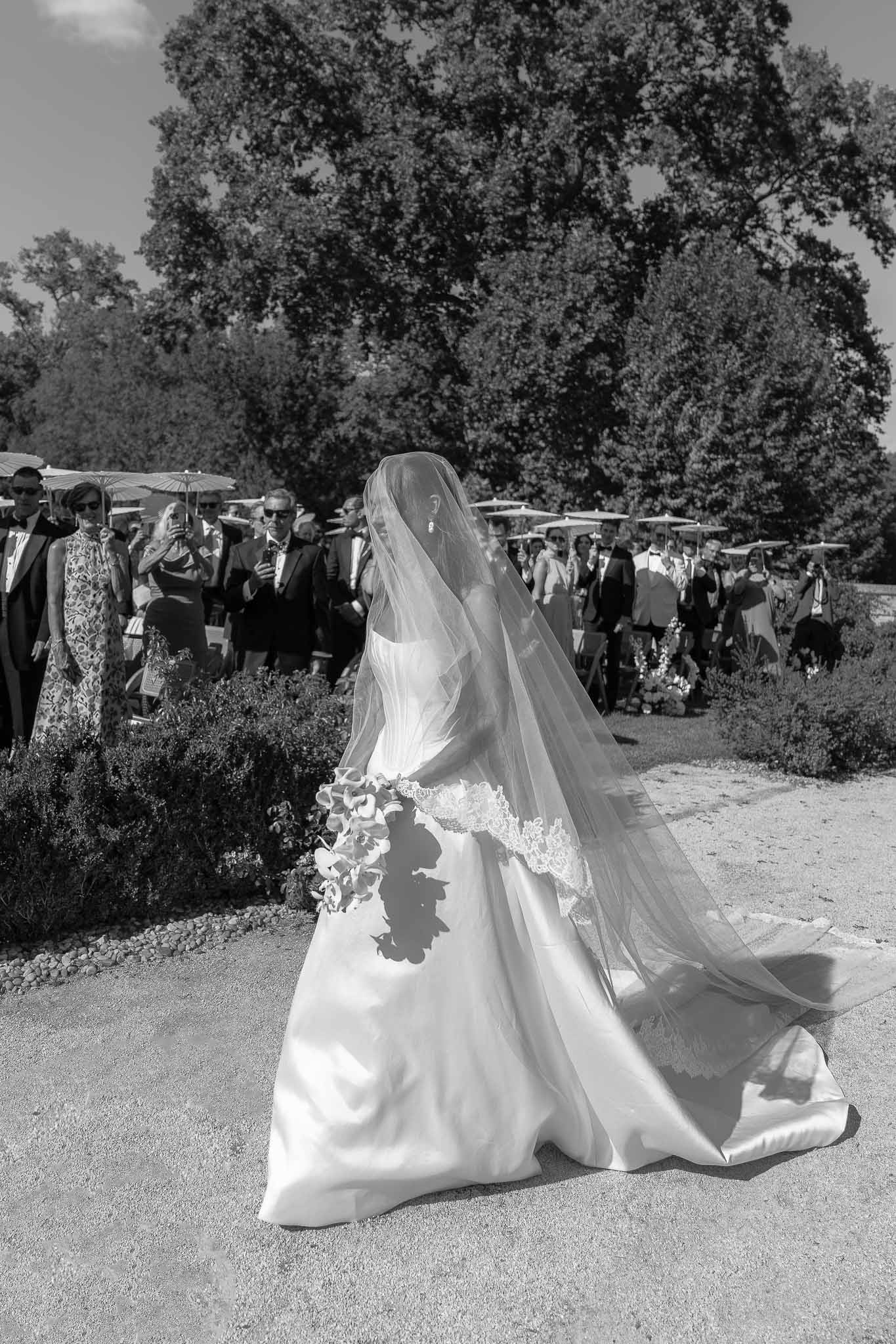 Black and white bride walking down gravel aisle with cathedral lace veil billowing toward seated guests