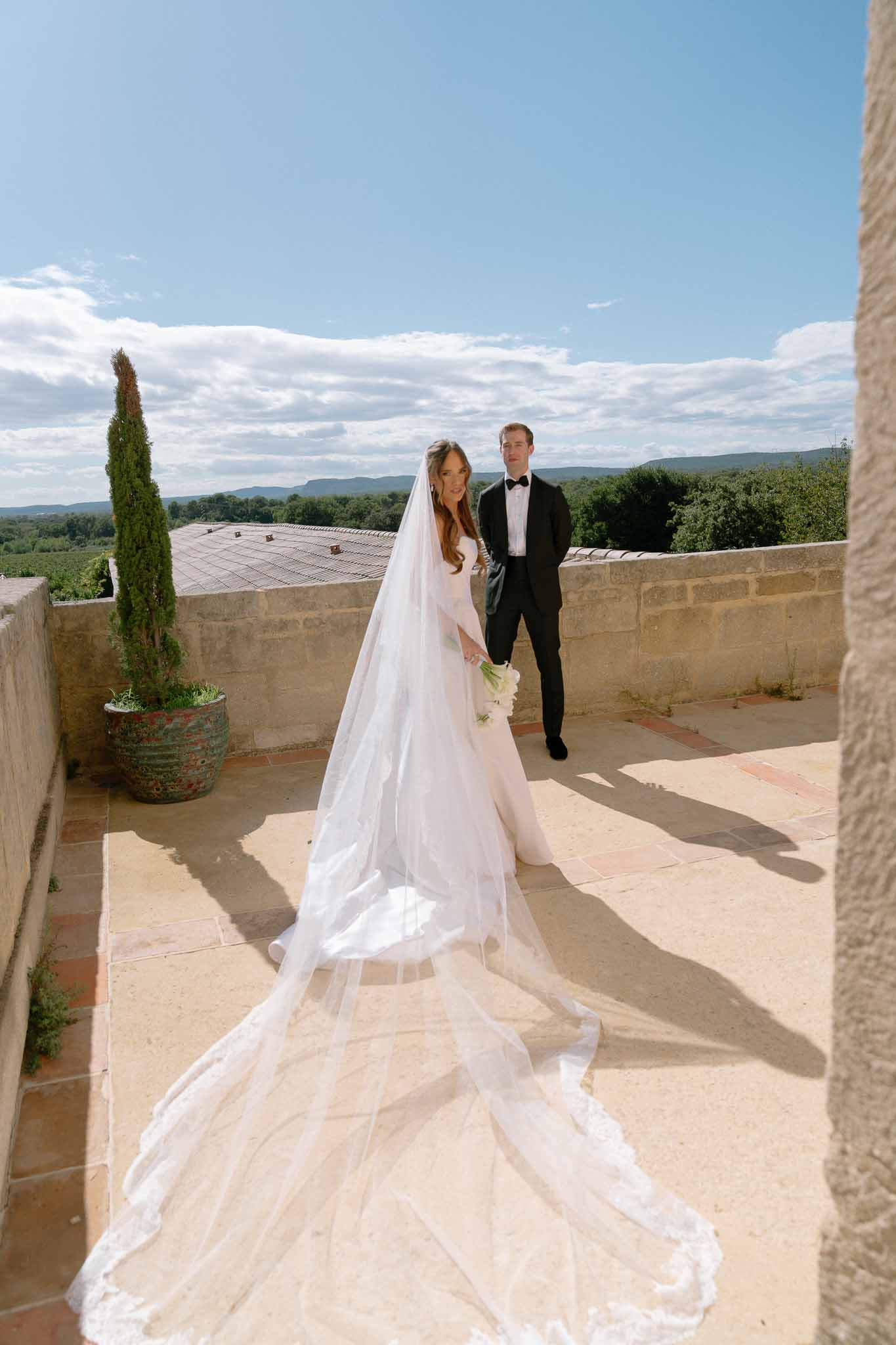 Bride in long-sleeve gown with lace-edged cathedral veil and groom in tuxedo on chateau terrace with vineyard view