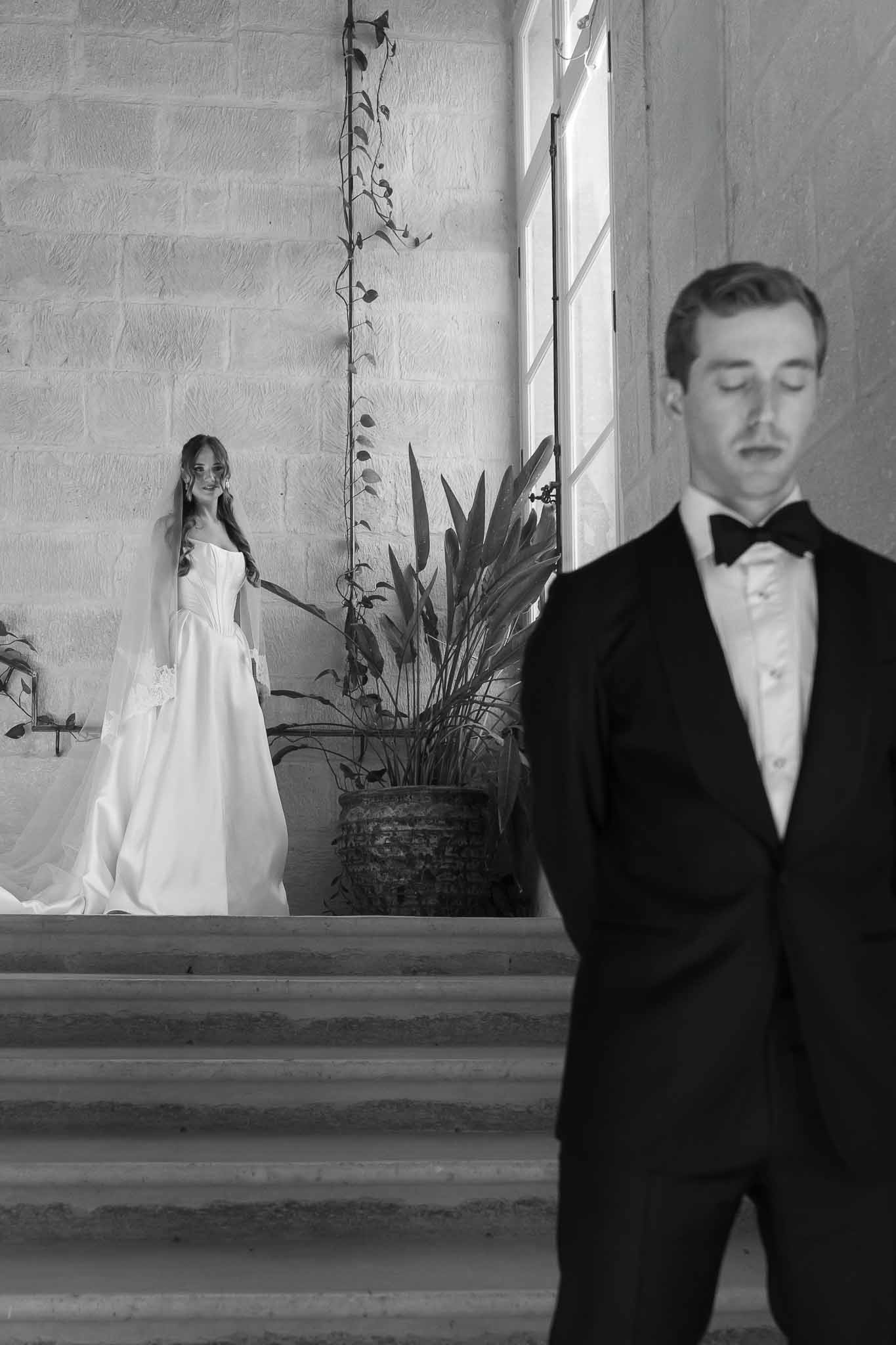 Black and white split-focus portrait with groom eyes closed on stone staircase and bride smiling in ballgown above