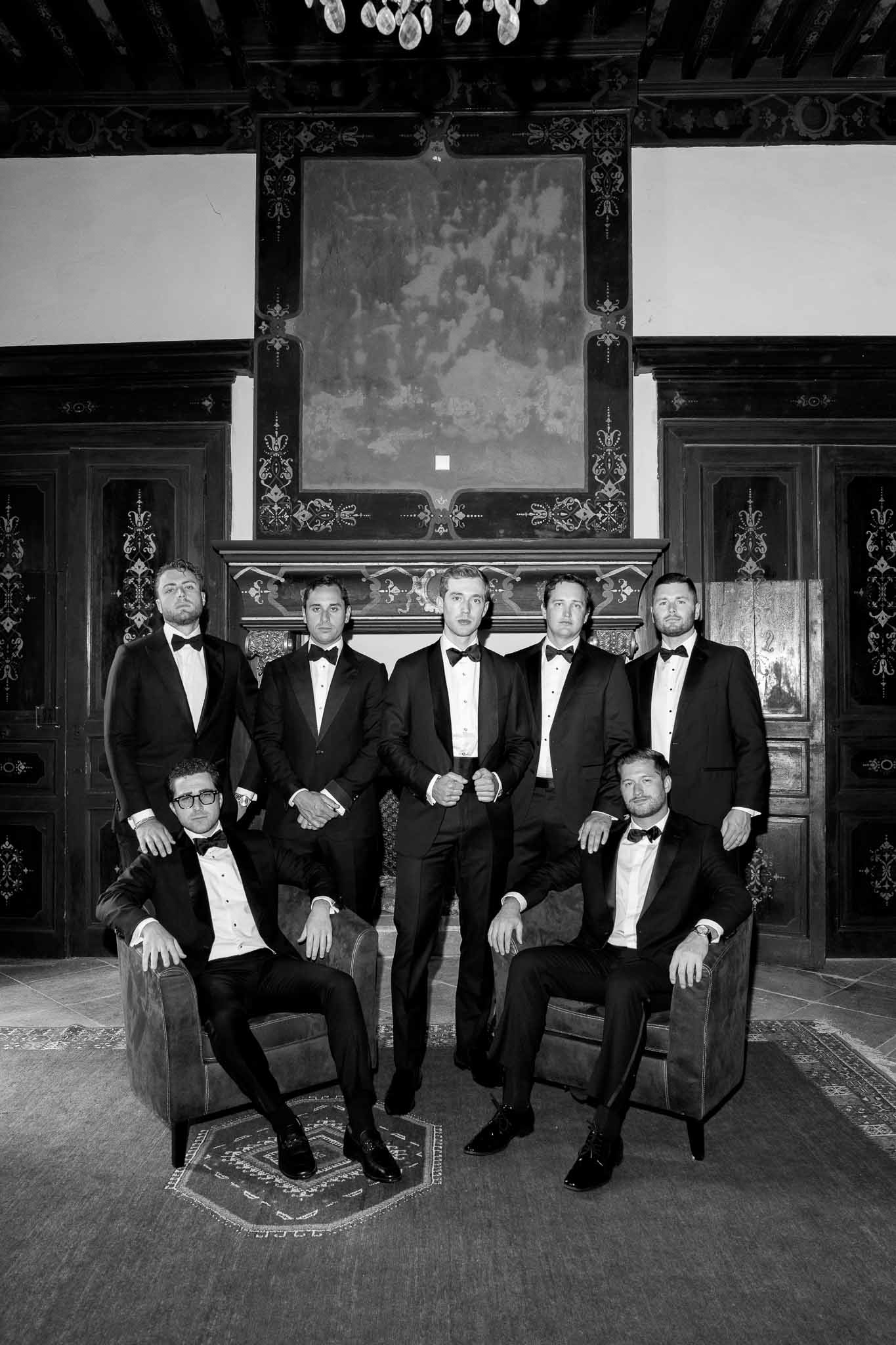 Black-and-white group portrait of groom and six groomsmen in black tuxedos posed in ornate chateau salon