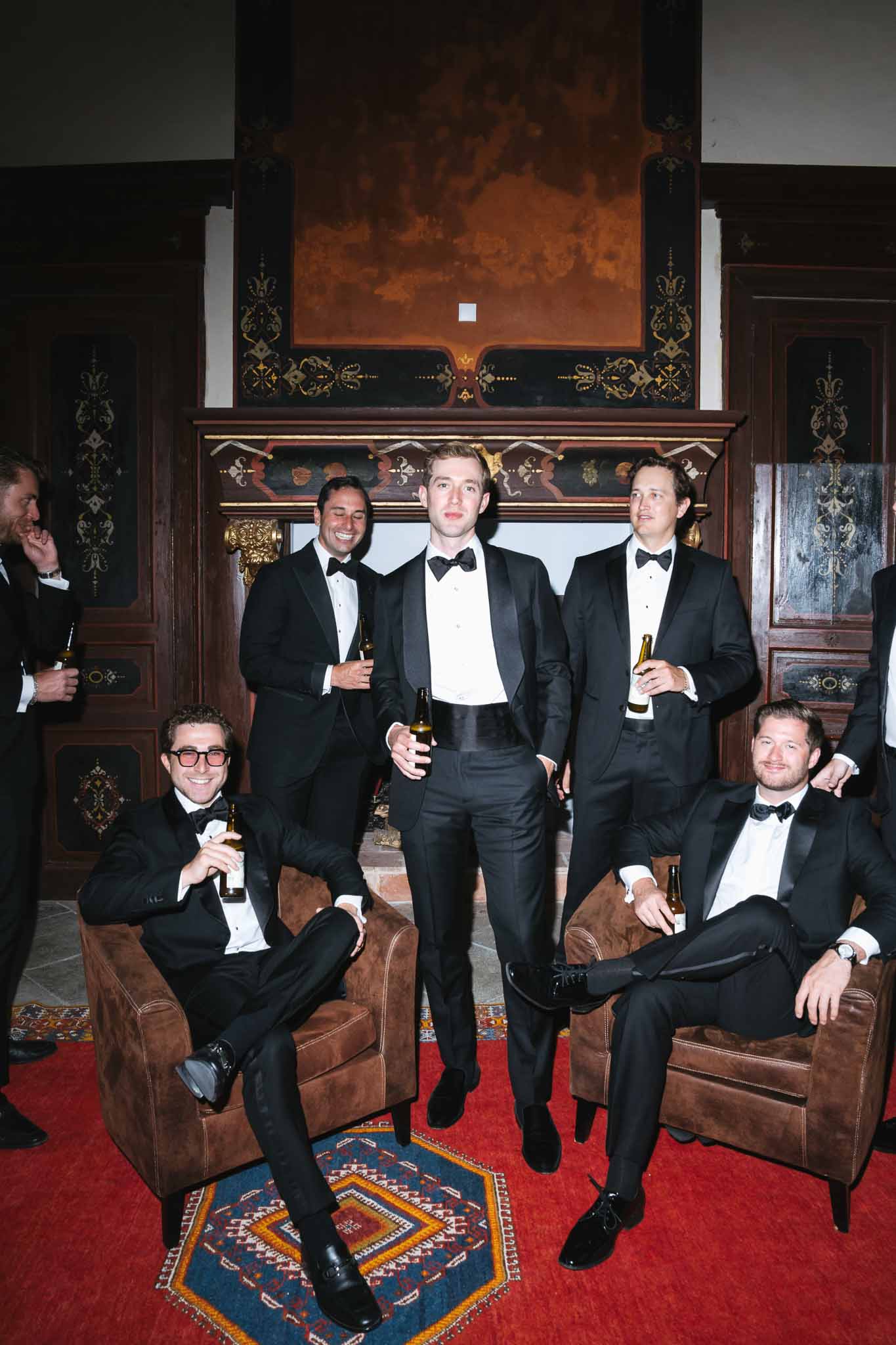 Groom and five groomsmen in tuxedos holding beer bottles in wood-panelled chateau room with painted fireplace and kilim rug