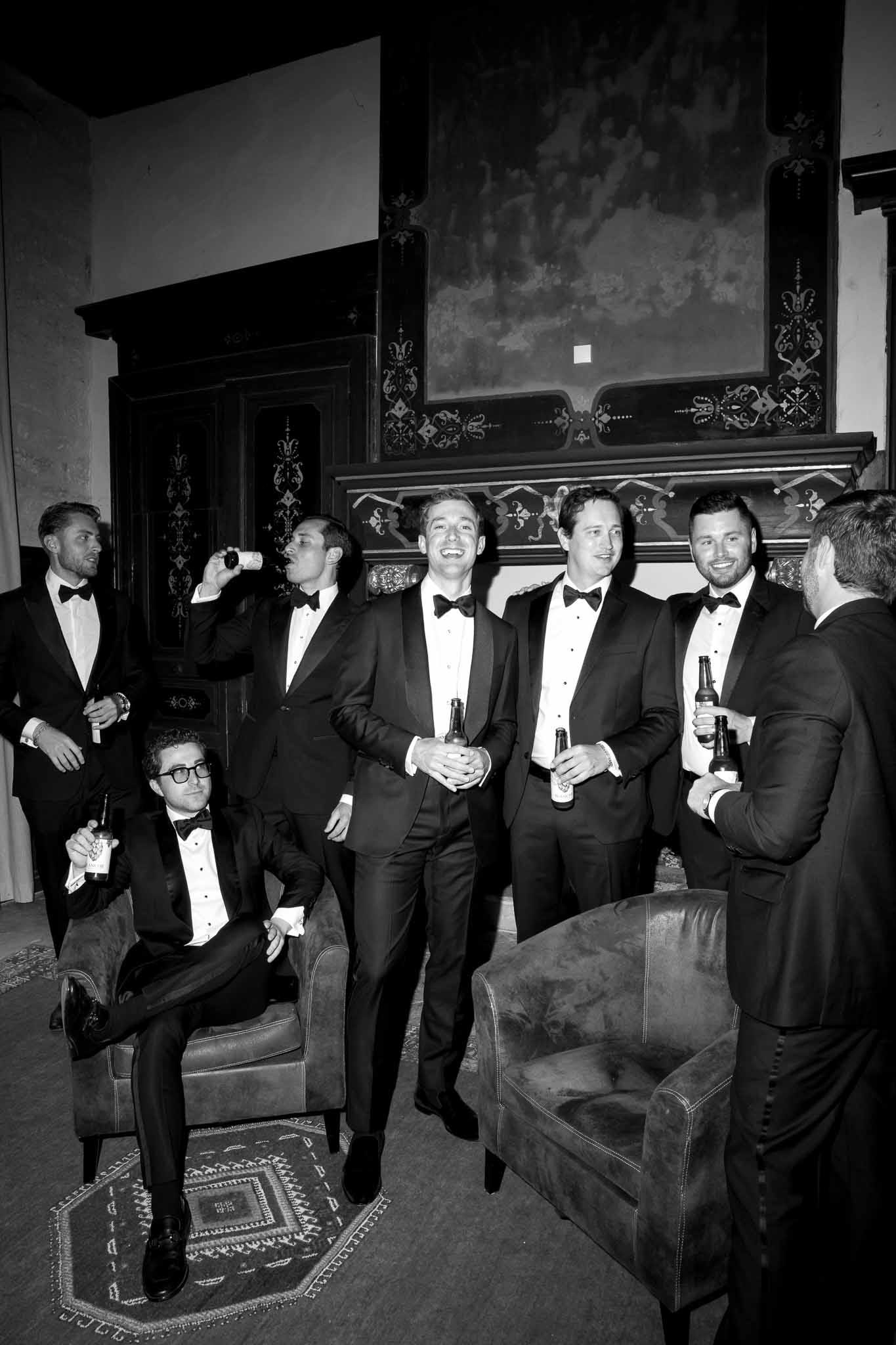 Black and white of seven groomsmen in tuxedos relaxing with beers in ornate chateau fireplace room