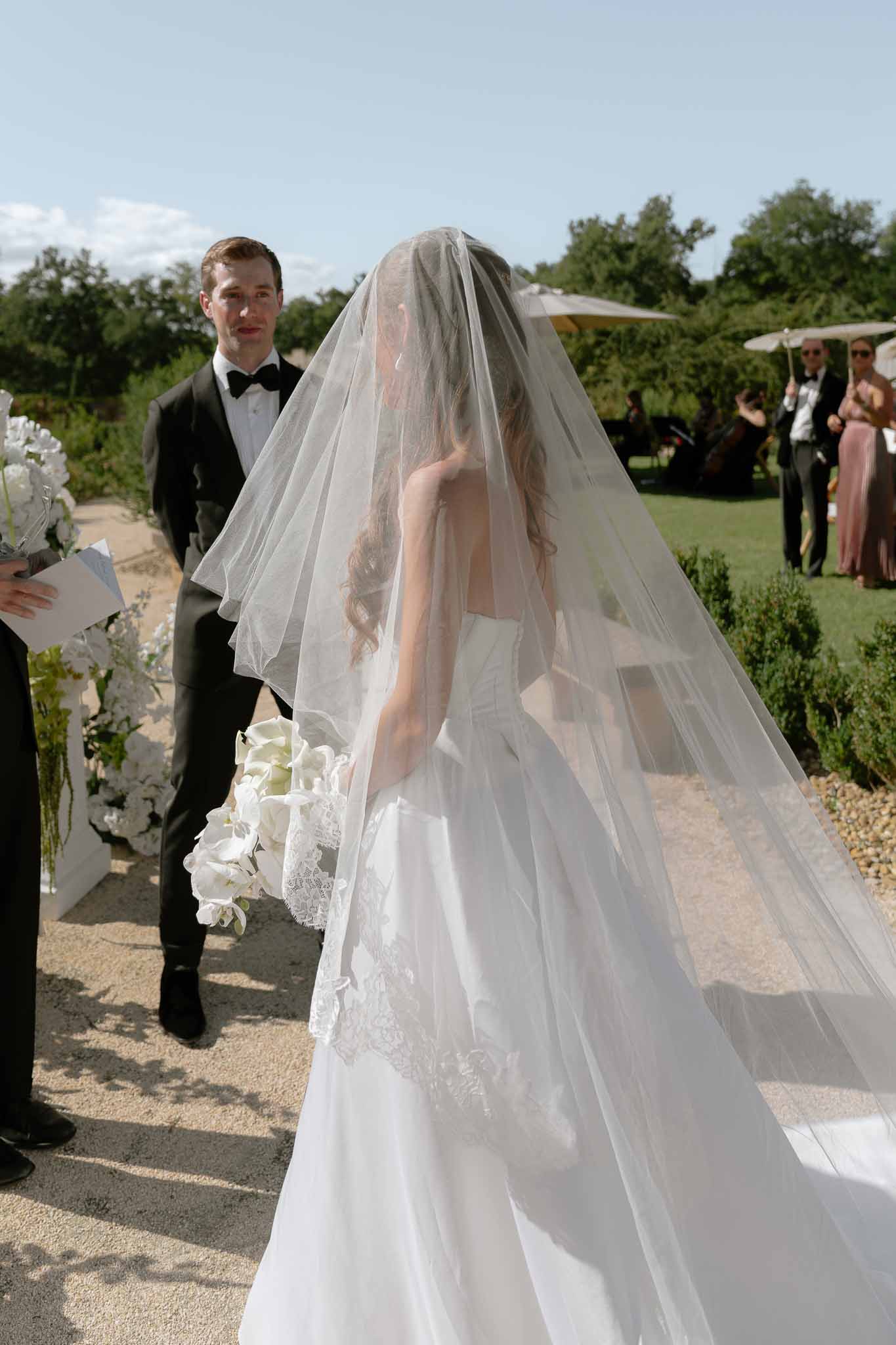 Bride in cathedral veil with orchid bouquet walks toward groom beside white floral arch in garden