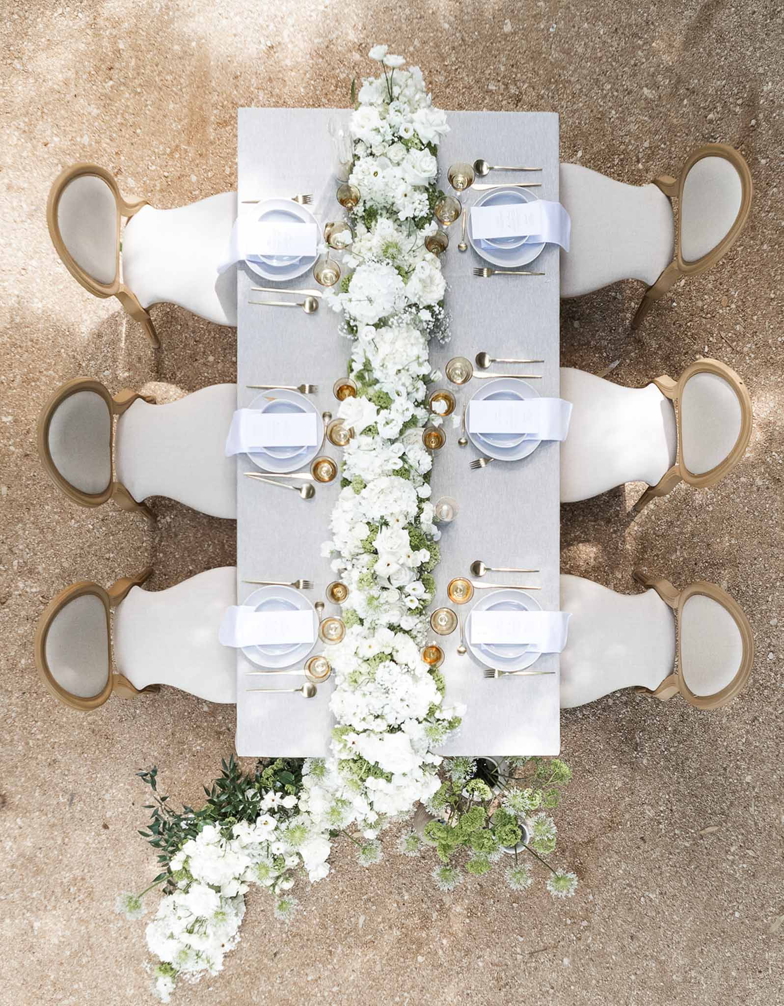 Aerial reception table with white hydrangea and ranunculus runner gold chairs amber glassware and overflowing ground florals