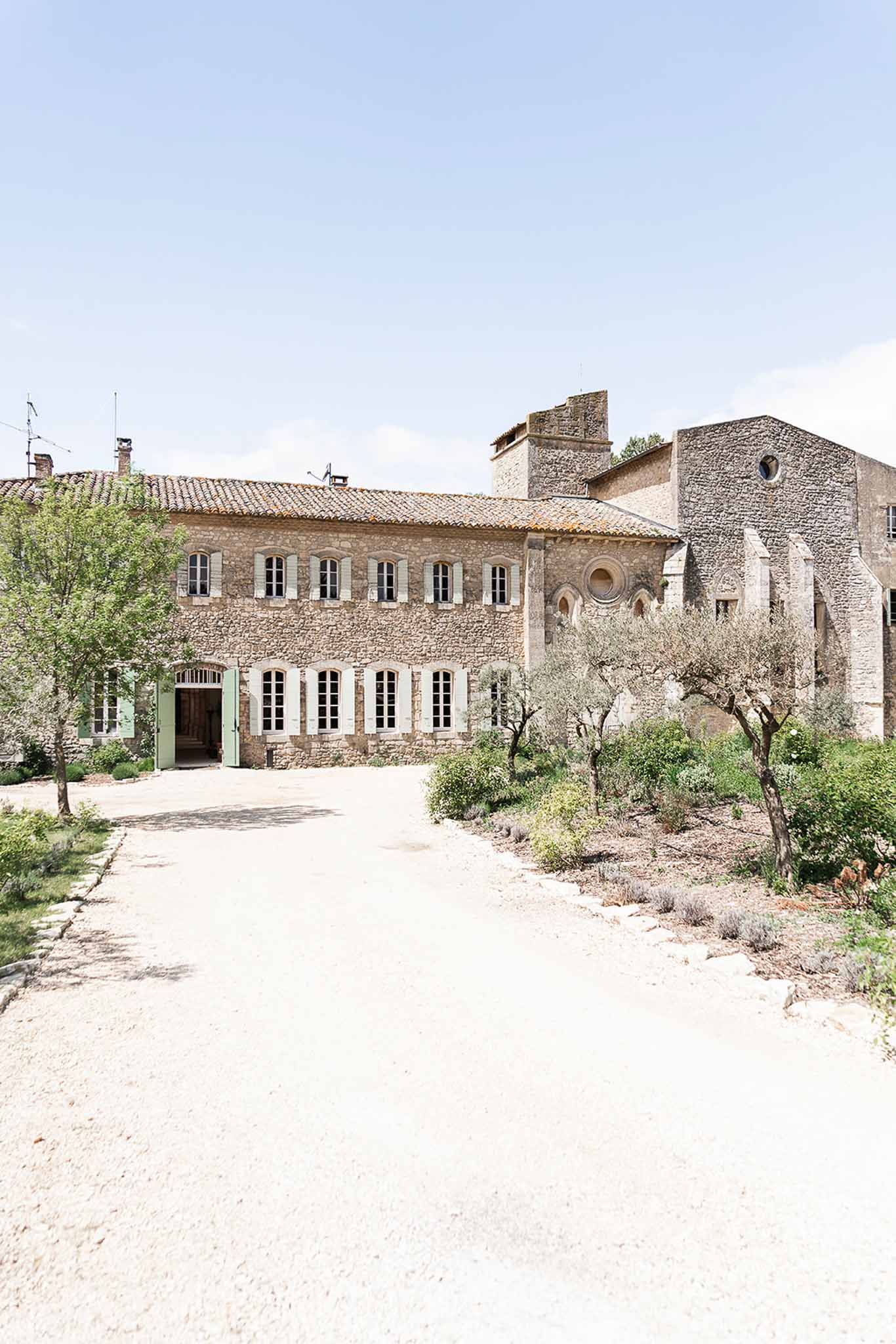 Stone mas with sage shutters beside Romanesque chapel with rose window on olive-lined gravel courtyard