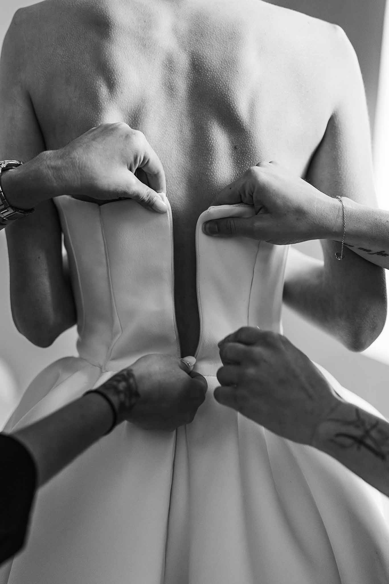 Black and white close-up of tattooed hands fastening corset-style wedding dress zipper on bride's back