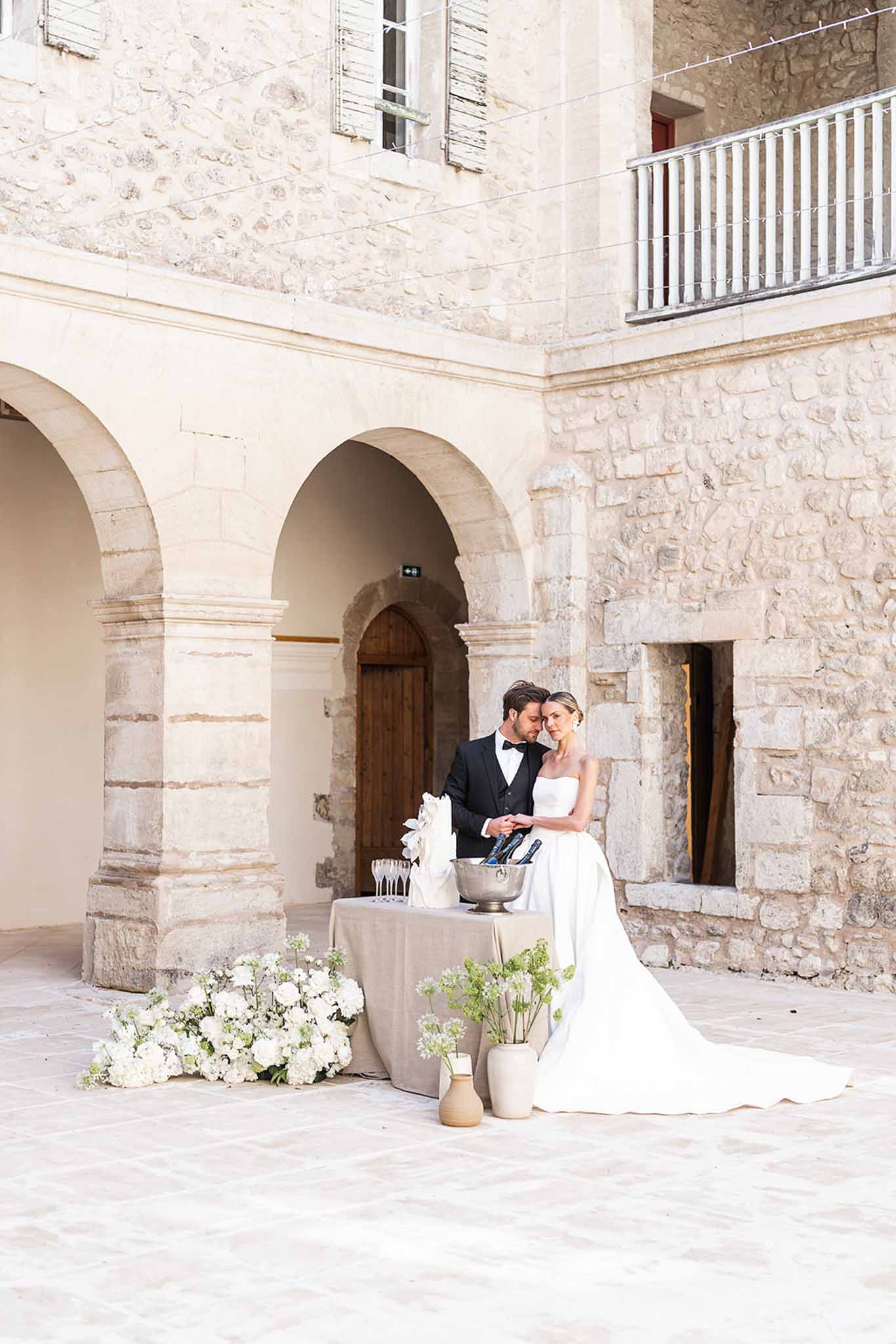 Bride and groom cutting white floral wedding cake in stone courtyard with arched colonnades and champagne flutes