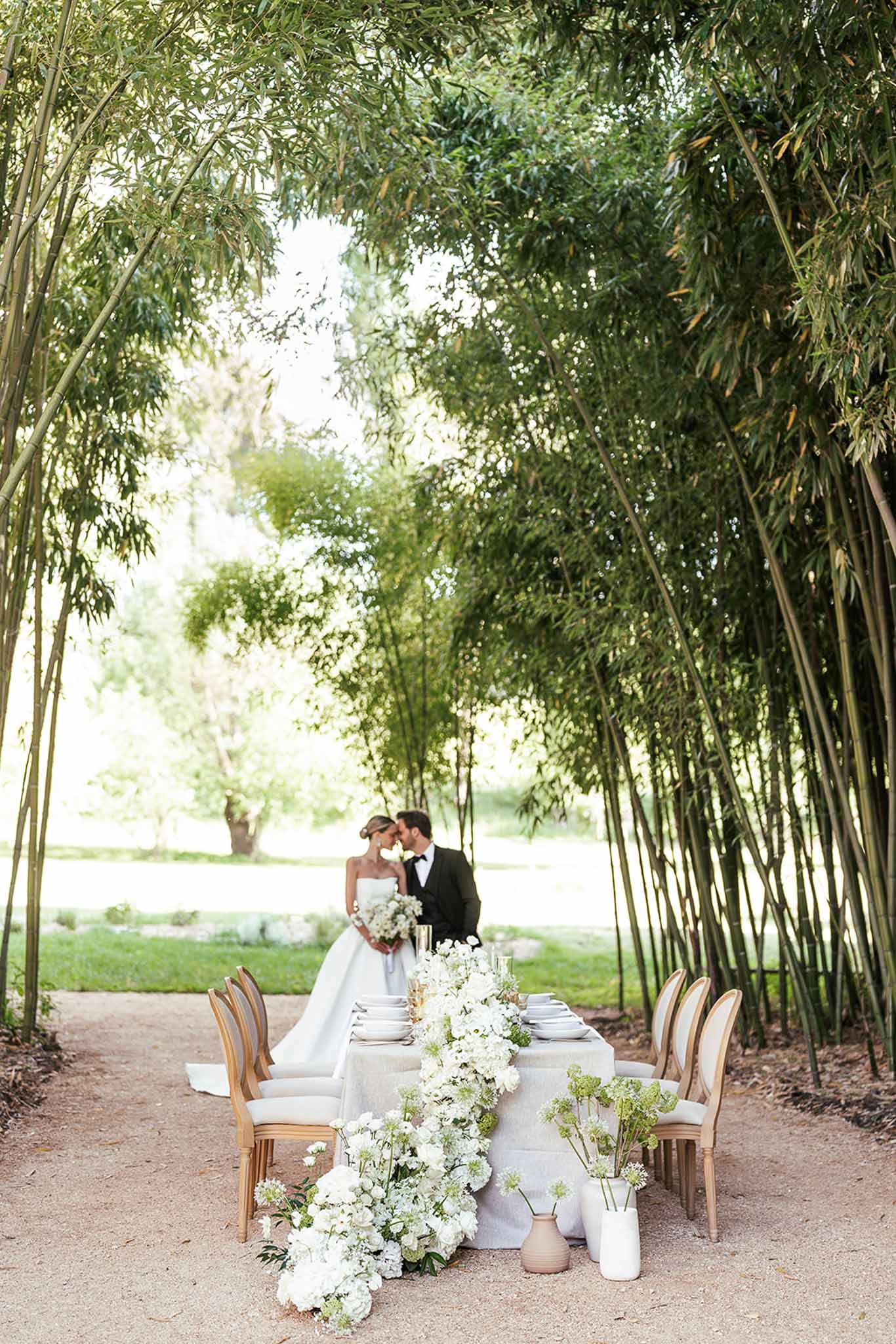 Couple behind grey linen table with cascading white hydrangea and ranunculus runner and carved wood chairs in garden