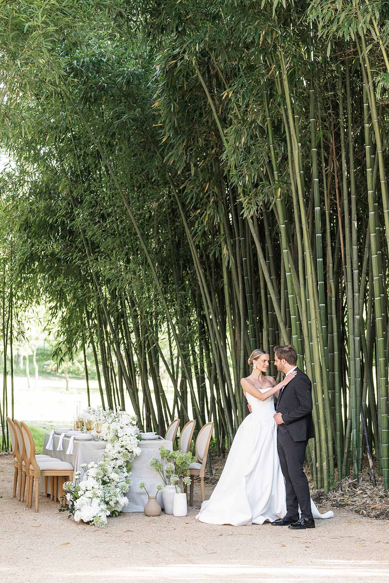 Couple before bamboo grove beside grey linen table with white hydrangea runner and gold glassware