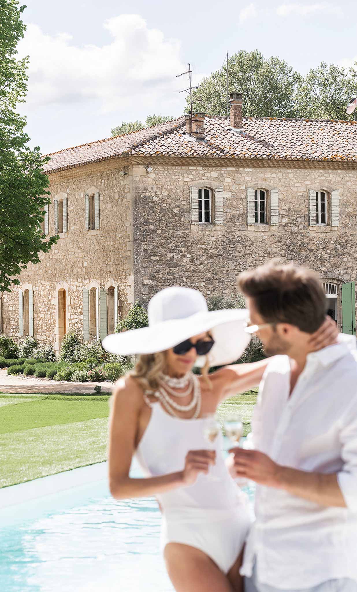 Couple holding champagne flutes by pool with French stone manor and manicured lawn in background