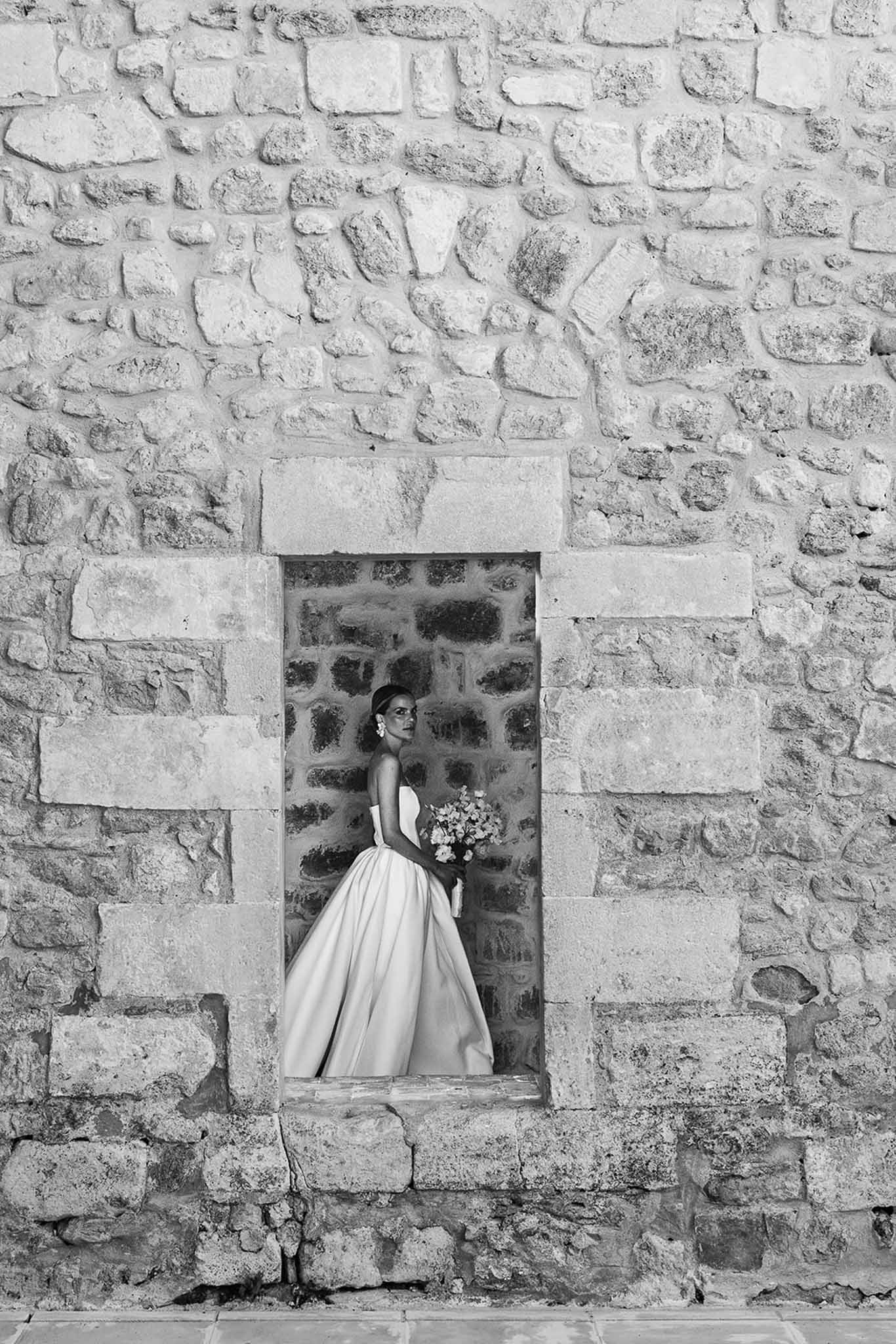 Black and white of bride in strapless ballgown standing in stone window opening with bouquet and headband
