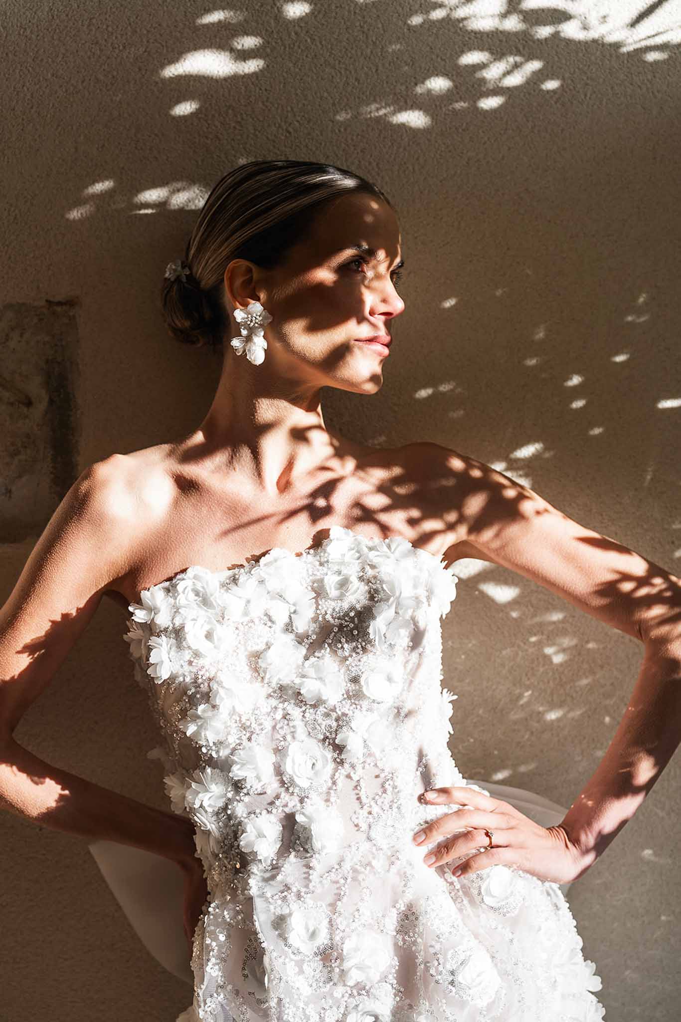 Bride in sculptural strapless gown with 3D fabric flowers posing against sunlit wall with dappled leaf shadows