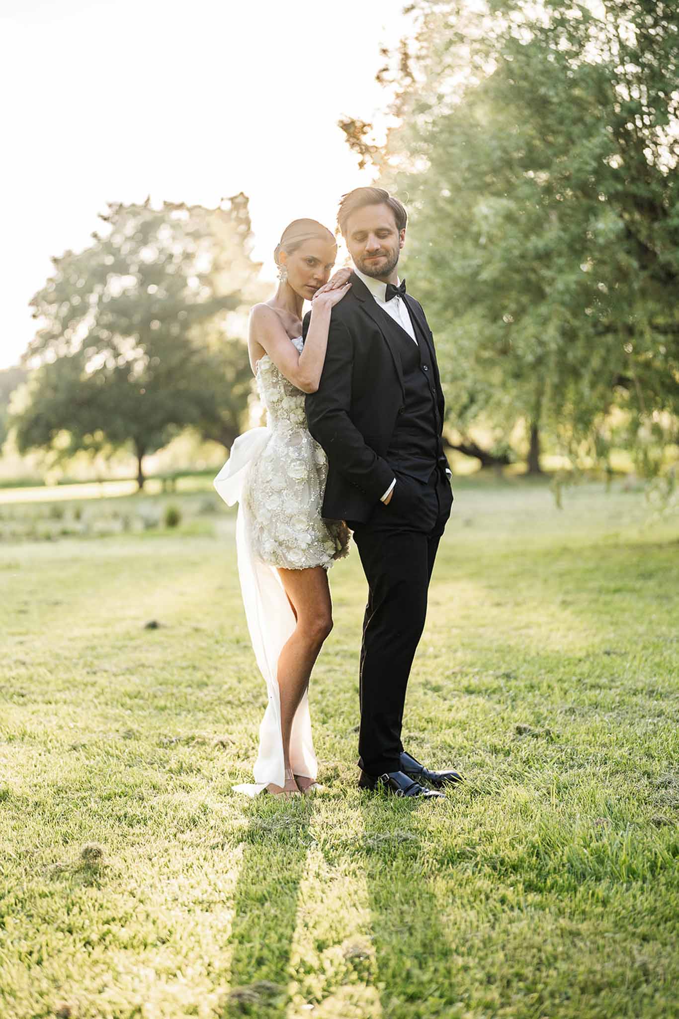 Bride in short strapless floral applique dress and groom in black tuxedo posing at golden hour in parkland