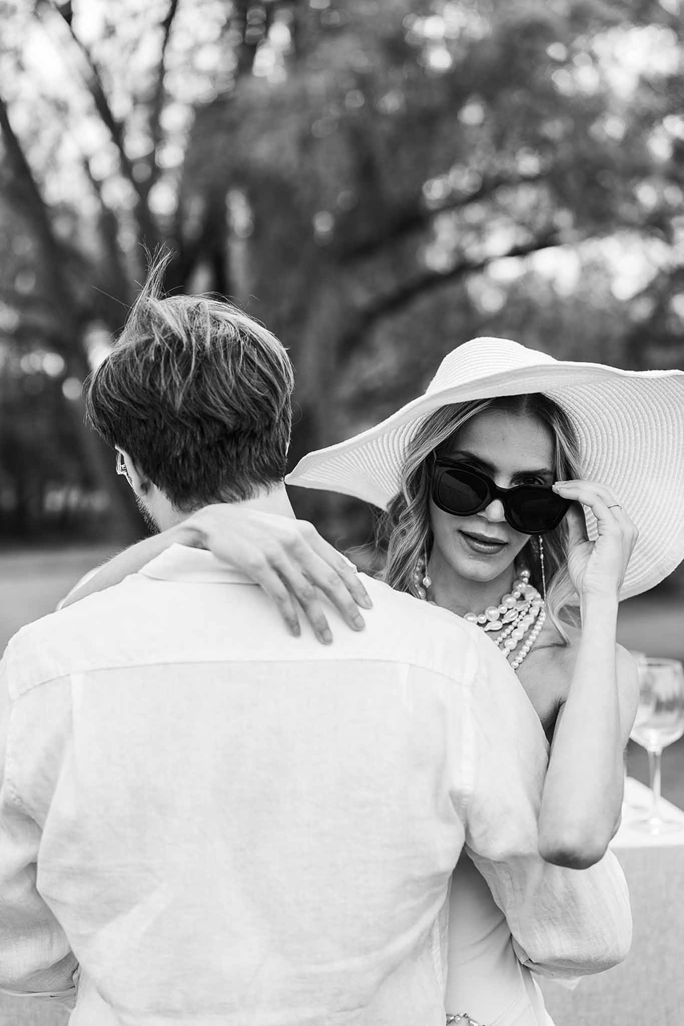 Black and white close-up of bride in sun hat and pearl necklace with groom at outdoor celebration