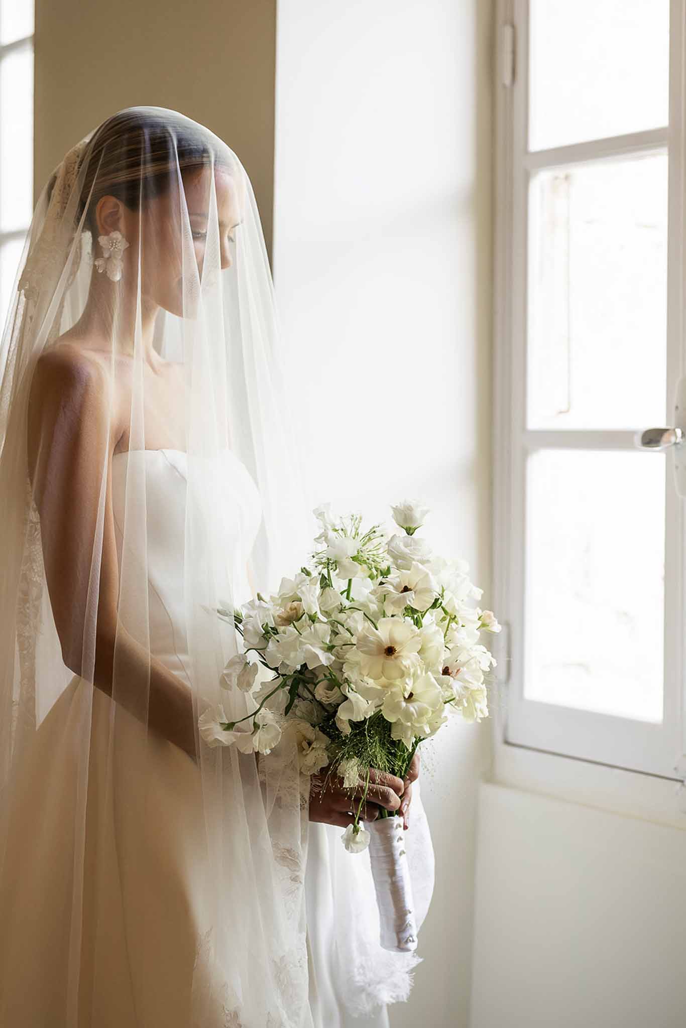 Bride in strapless gown with sheer veil holding all-white cosmos and sweet pea bouquet at tall window