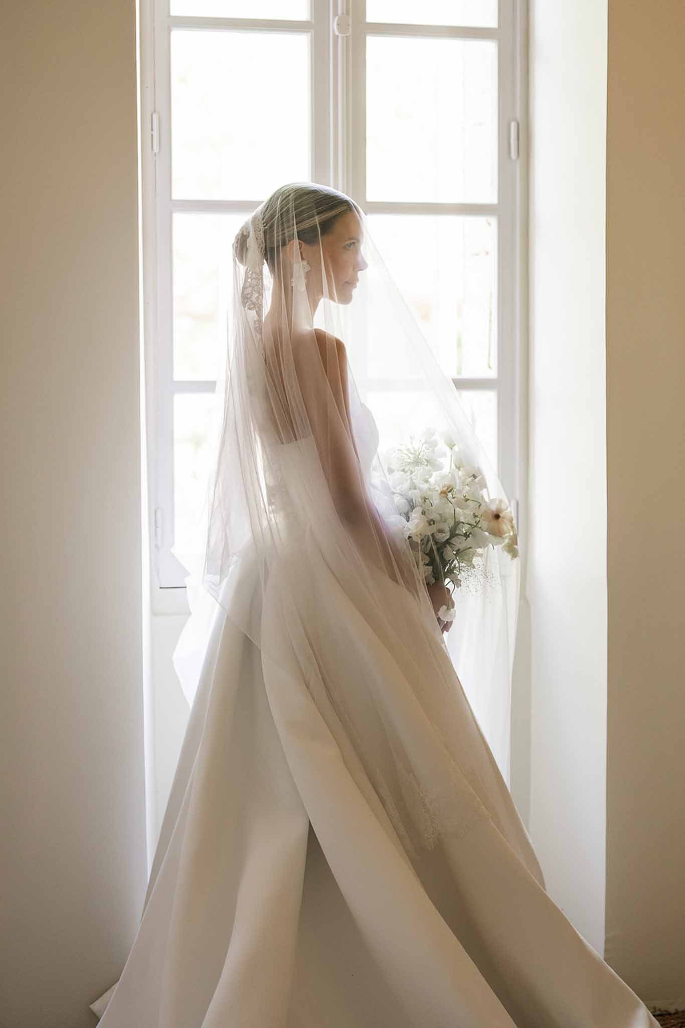 Bride silhouetted against backlit French window holding cosmos and sweet pea bouquet