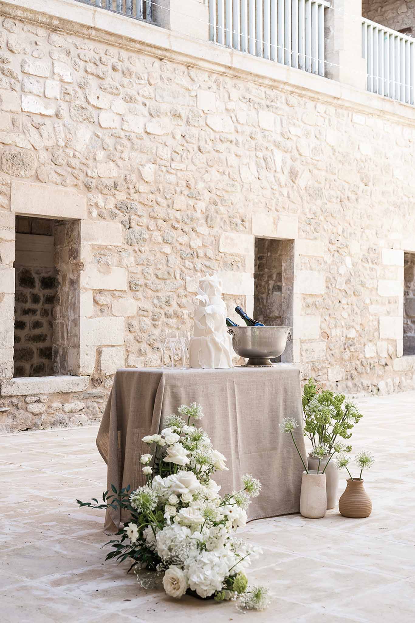 Wedding cake table with ruffled white cake, champagne, and ground-level florals in stone courtyard