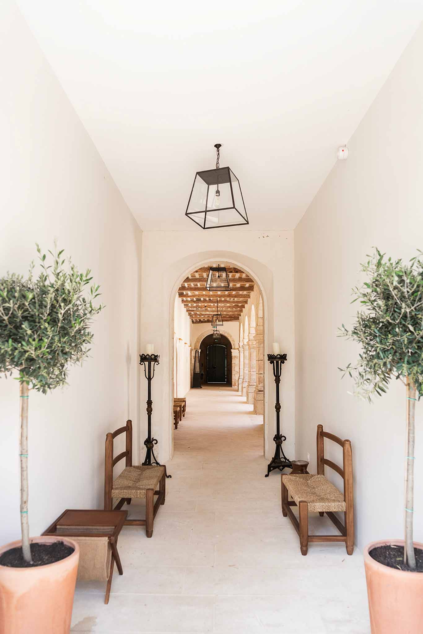 Chapel corridor with arched doorways, iron pendant lanterns, pillar candles, and potted olive trees