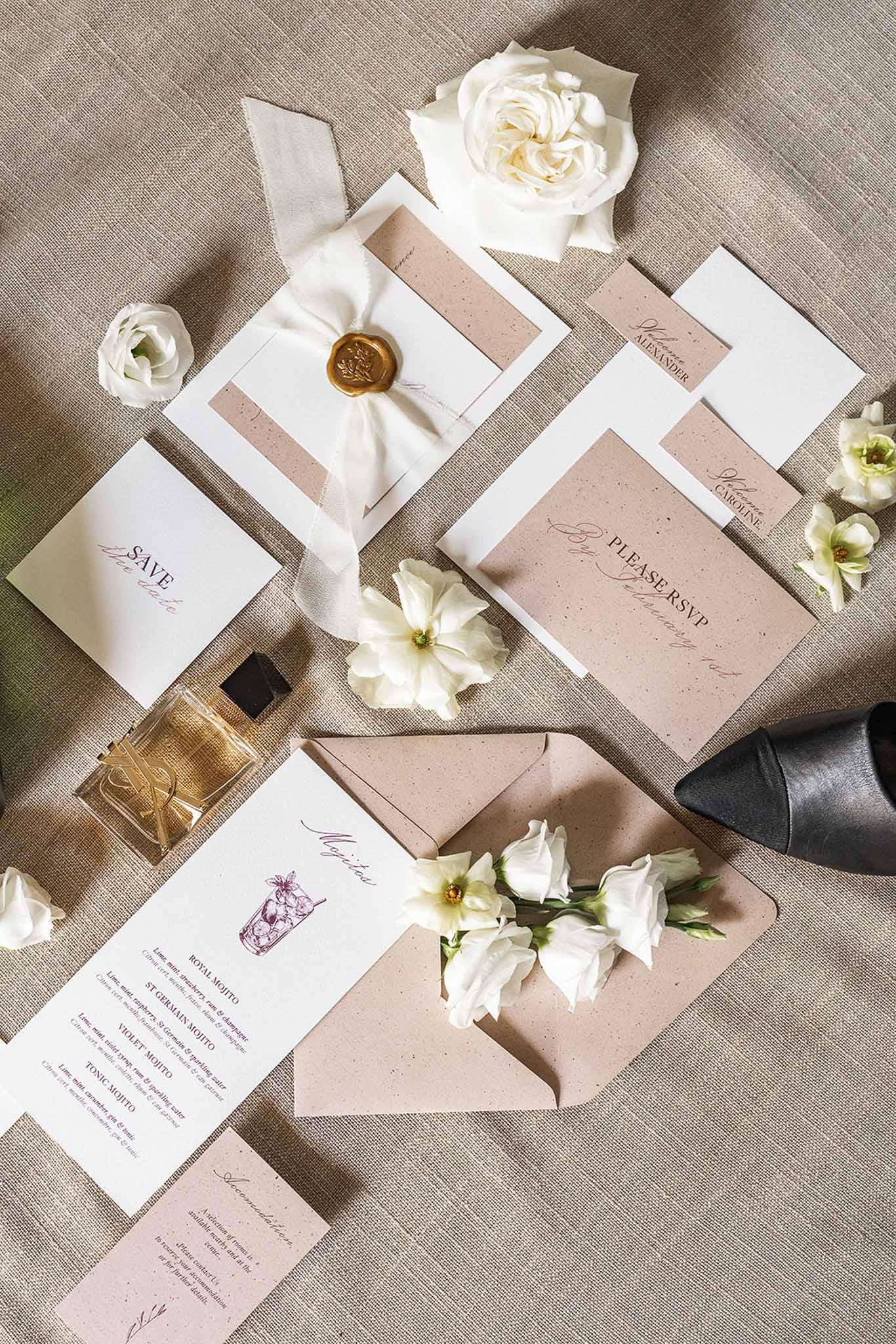 Stationery suite with gold wax seal, ivory ribbon, burgundy script, ivory roses, and perfume bottle on linen