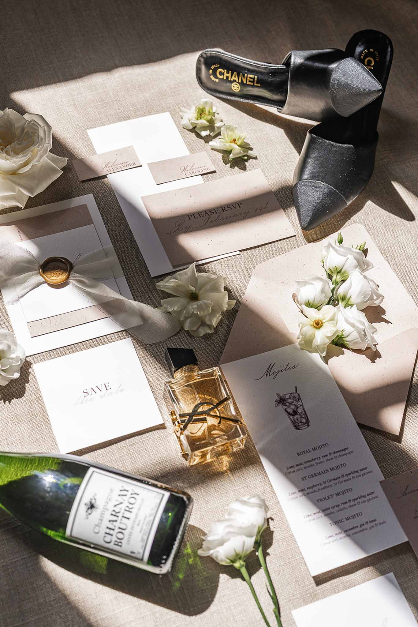 Flat lay of wedding stationery with gold wax seal, Chanel heels, YSL perfume, and champagne on natural linen