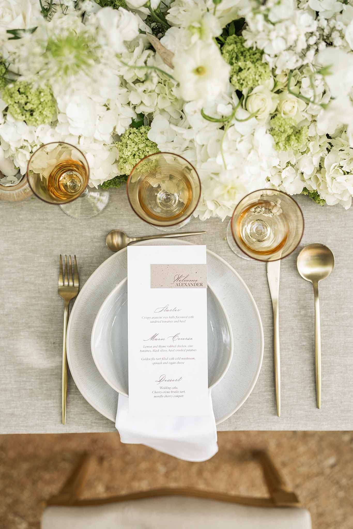 Overhead flat lay of reception place setting with grey plates, gold cutlery, menu card, and white hydrangea centerpiece