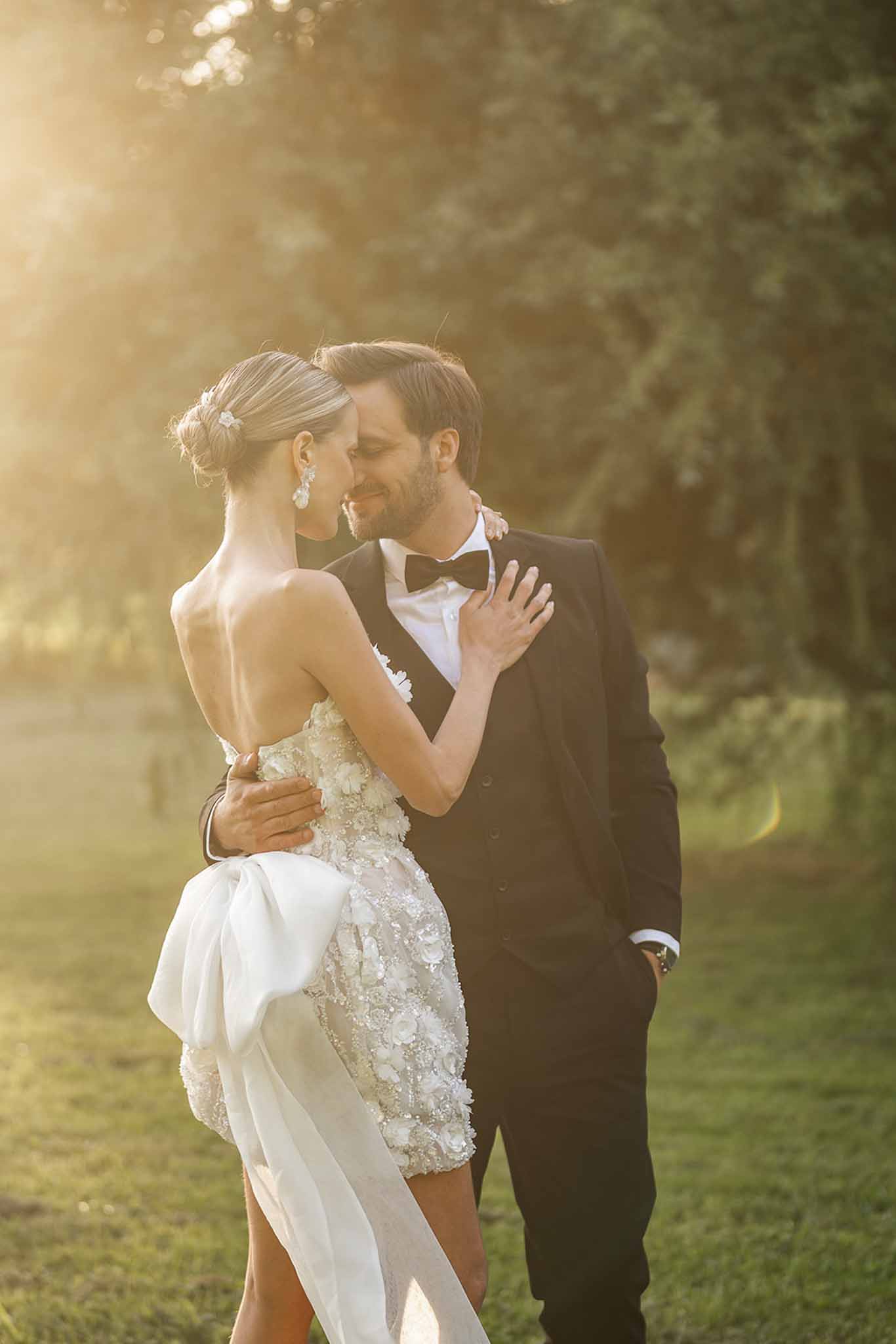 Bride and groom embrace on garden lawn at golden hour, bride in floral applique gown with bow detail