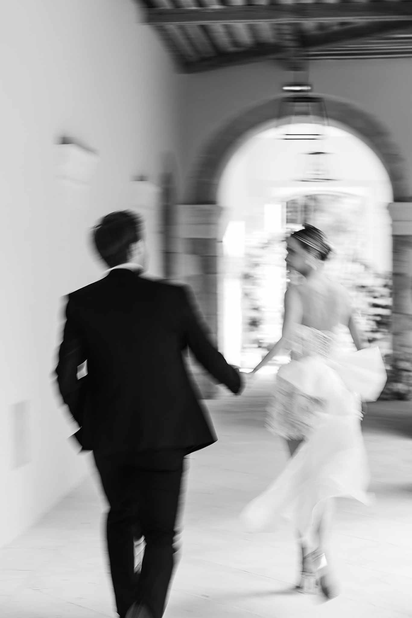 Black-and-white motion-blurred image of bride and groom running hand in hand through a chateau corridor