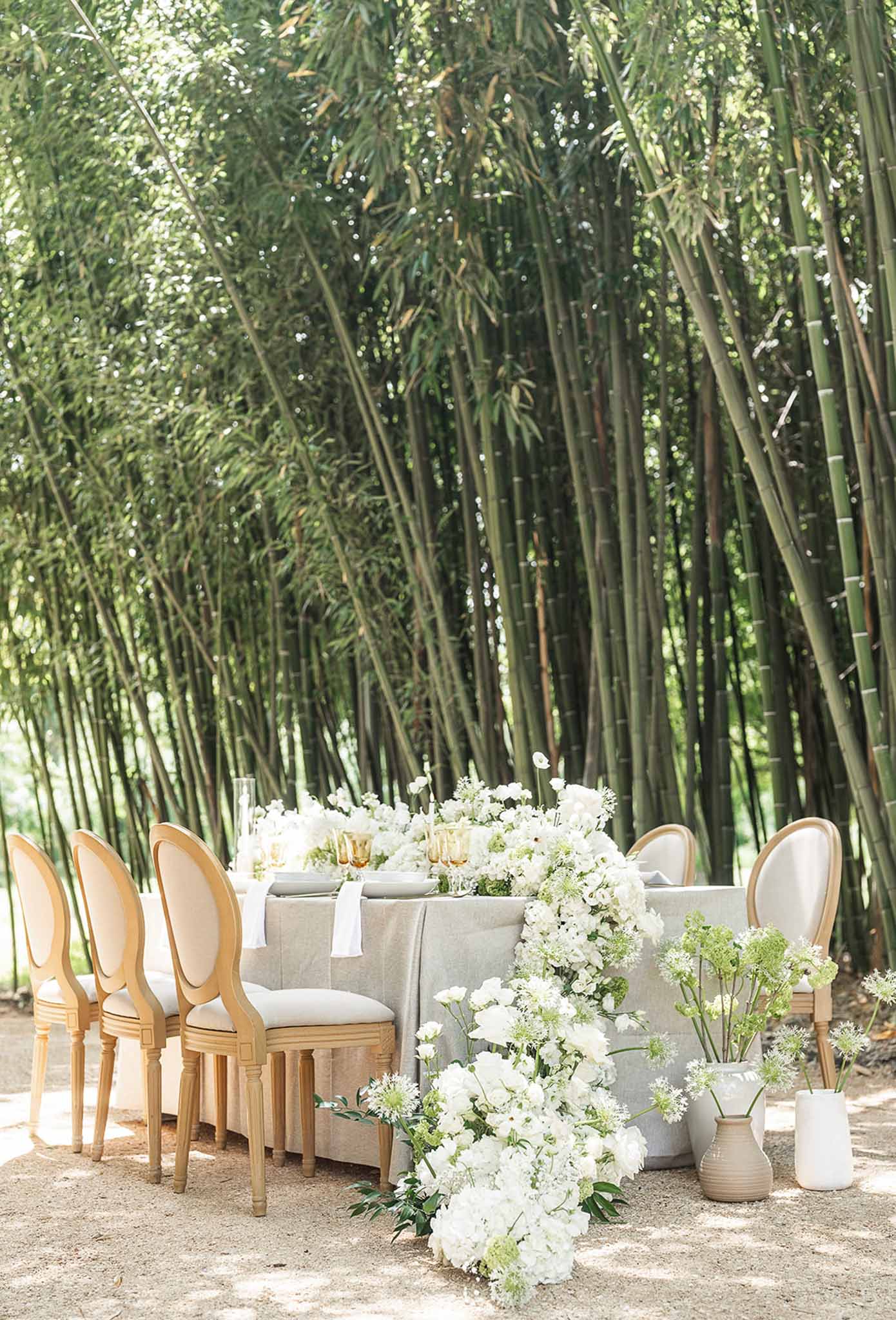 Editorial tablescape with white peony runner, amber glasses, and oval-back chairs before bamboo grove