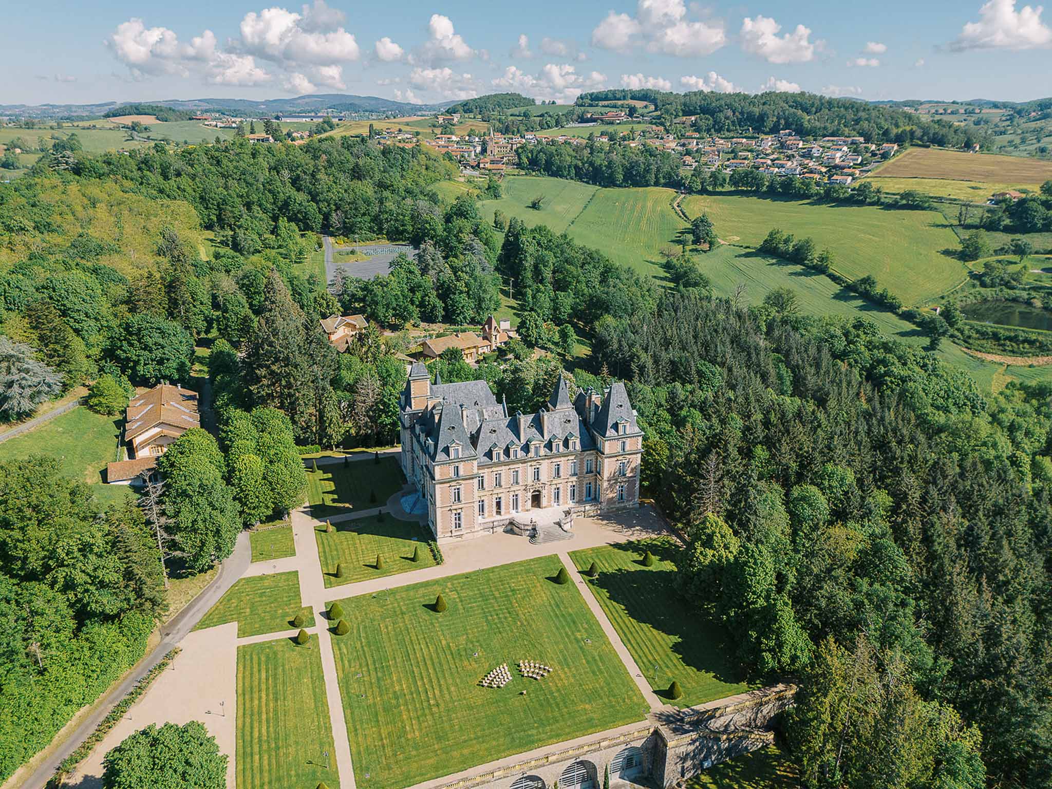 Aerial view of turreted chateau with parterre gardens, topiary, front lawn ceremony chairs, and farmland