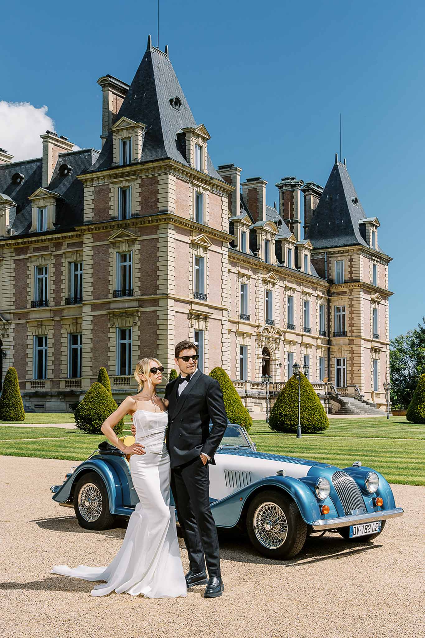 Bride and groom in sunglasses leaning against vintage Morgan roadster in front of Belle Epoque chateau