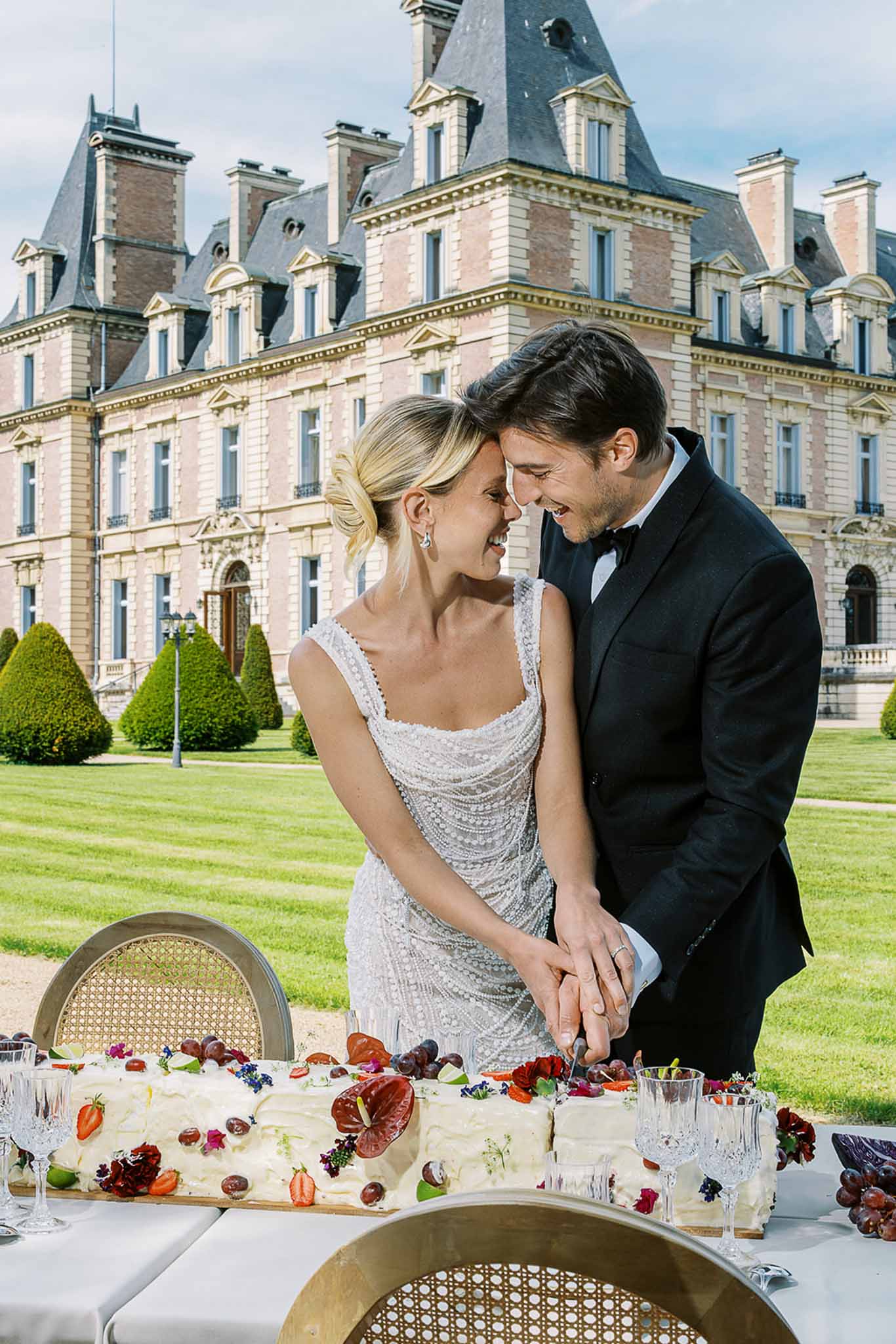 Laughing couple cutting rectangular berry and anthurium cake before grand chateau facade