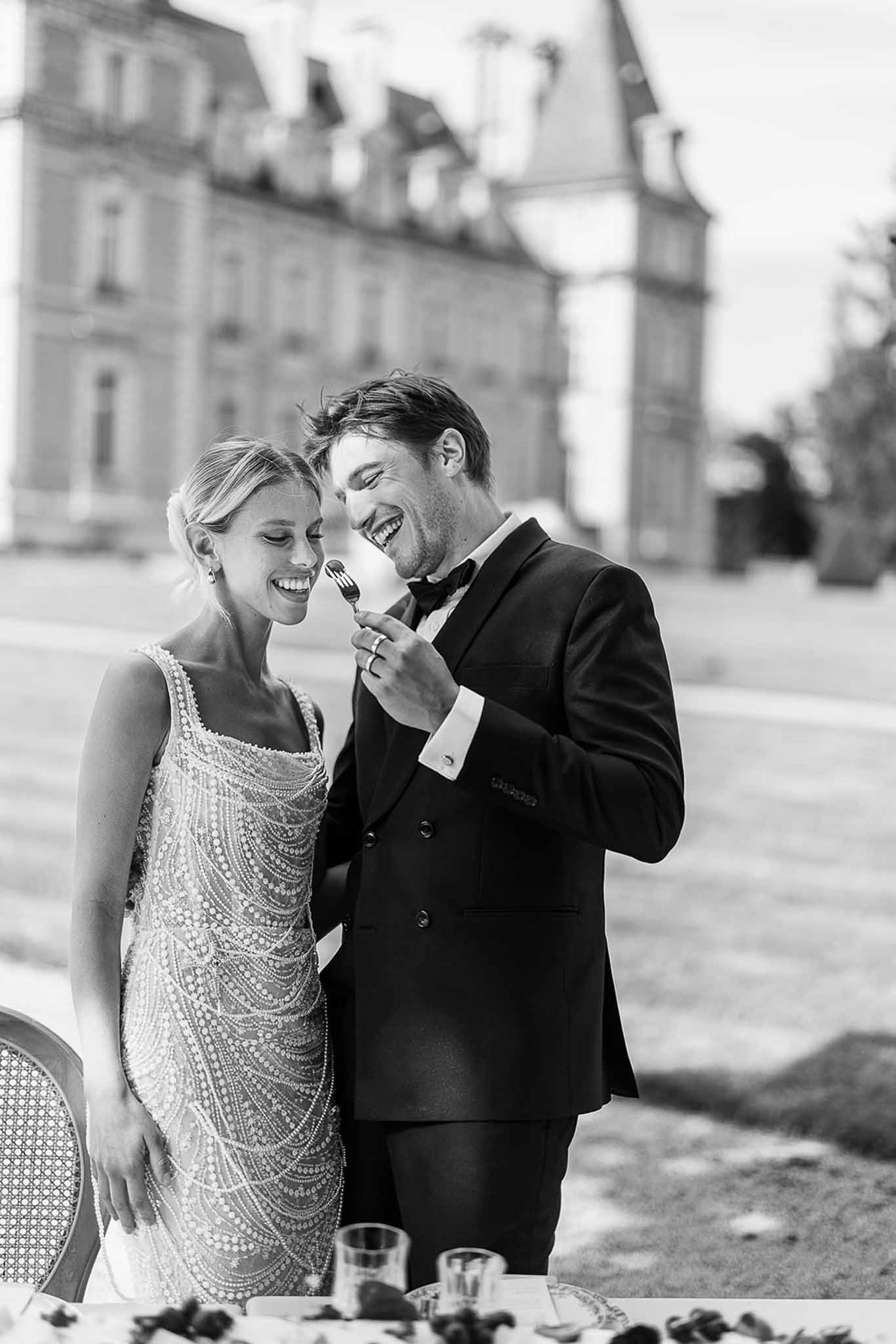 Black-and-white portrait of laughing couple sharing dessert on chateau grounds, bride in beaded mini dress