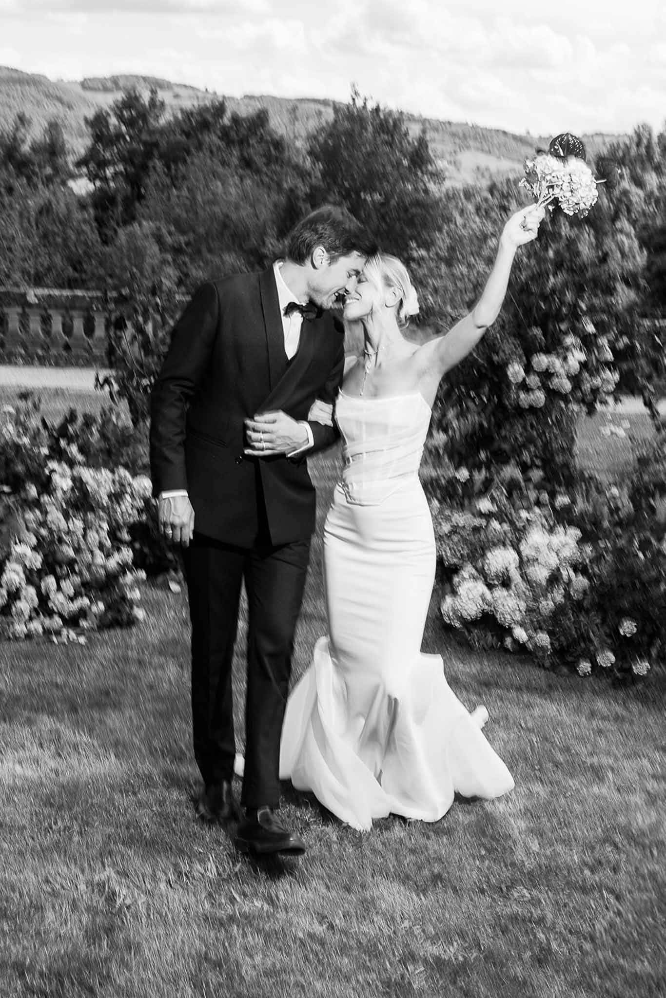 Black and white of couple laughing in rain with bouquet raised overhead before circular hydrangea floral installation