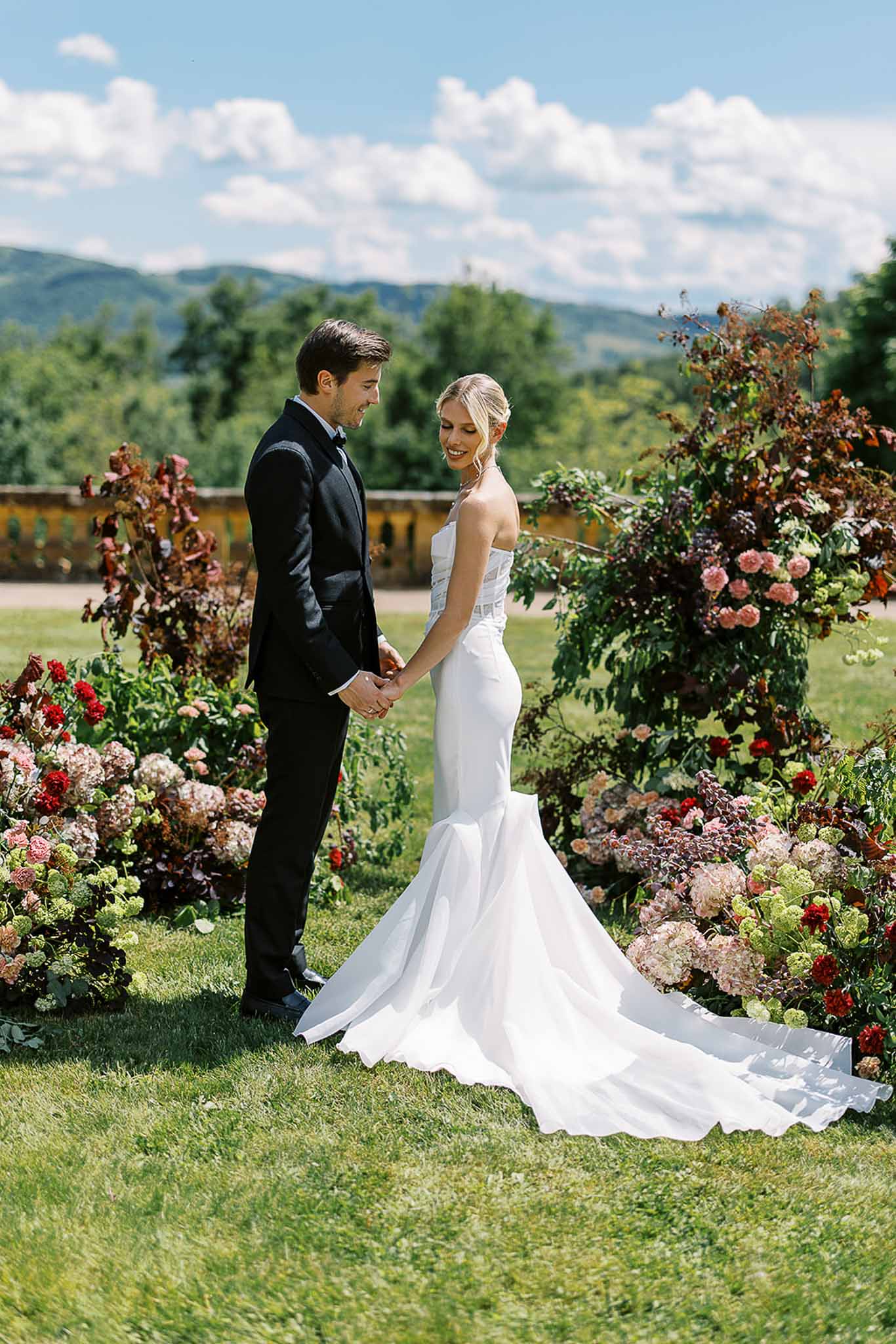 Bride and groom holding hands on a lawn in front of a burgundy and blush ground-level floral installation