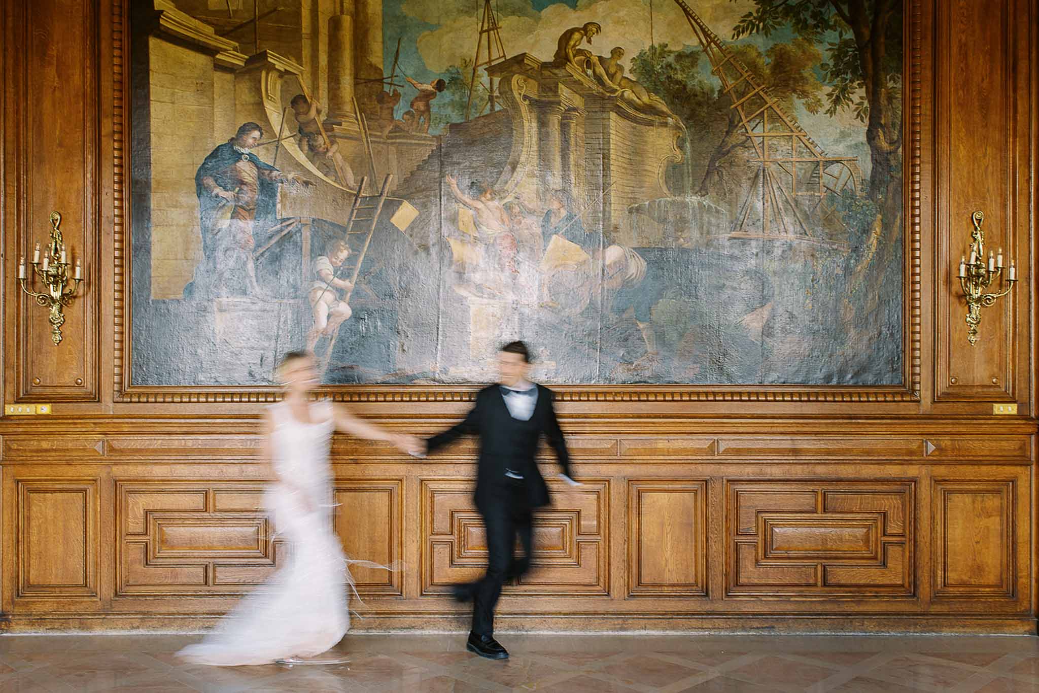 Motion-blurred couple running through oak-panelled hall with gold sconces and antique oil painting