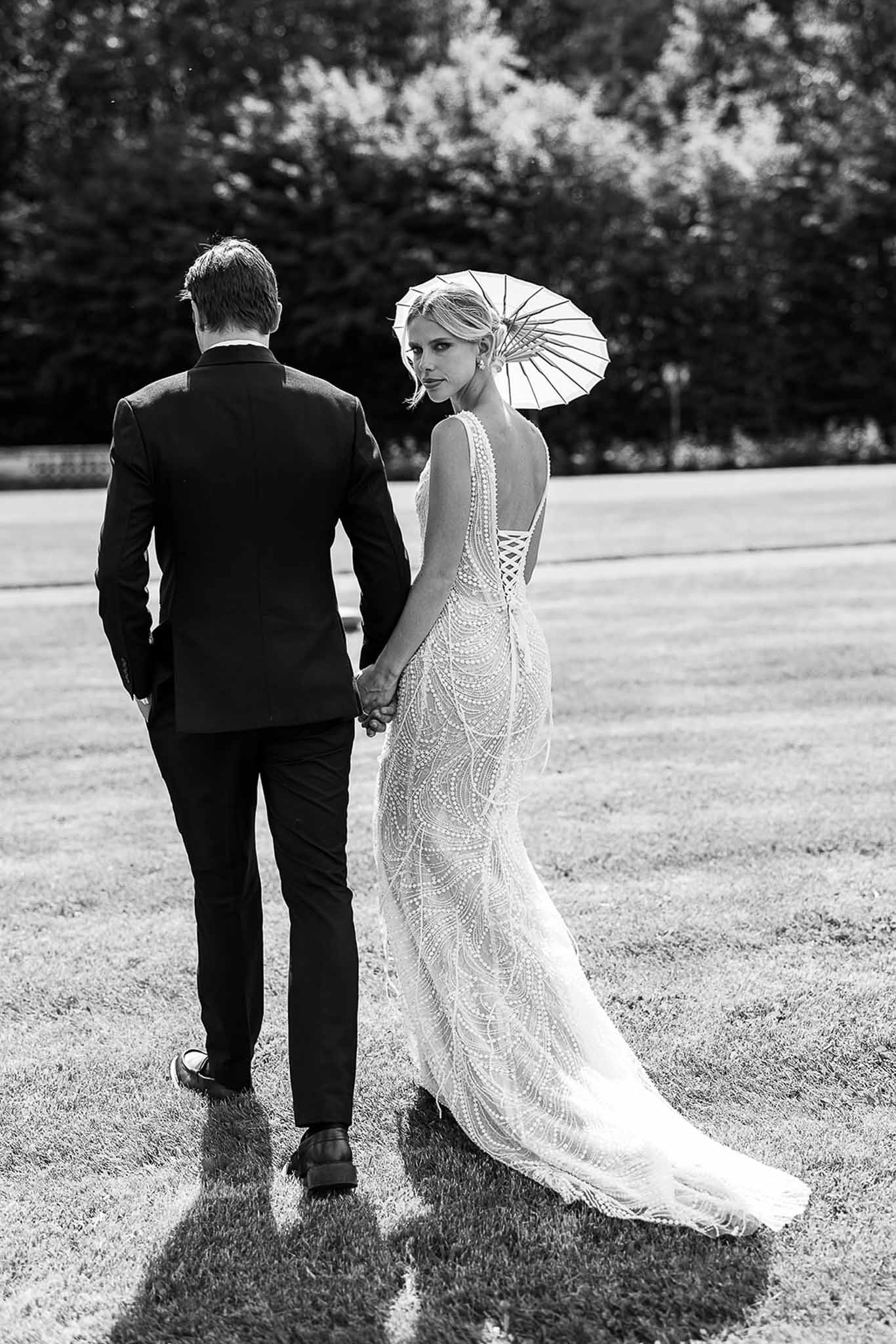 Black-and-white portrait of couple walking away on lawn as bride looks back holding paper parasol