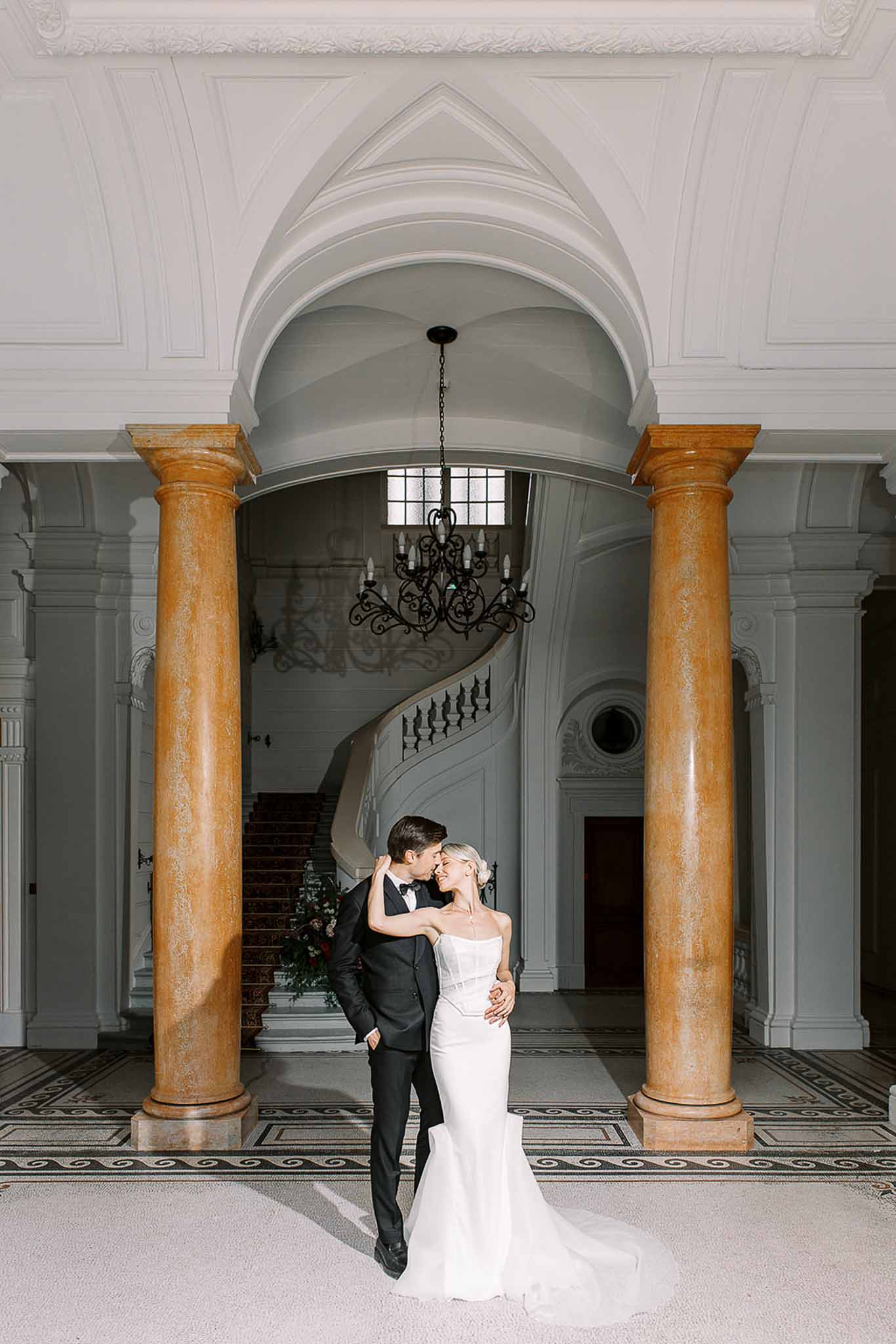 Couple foreheads touching between marble columns with curved staircase and mosaic floor behind