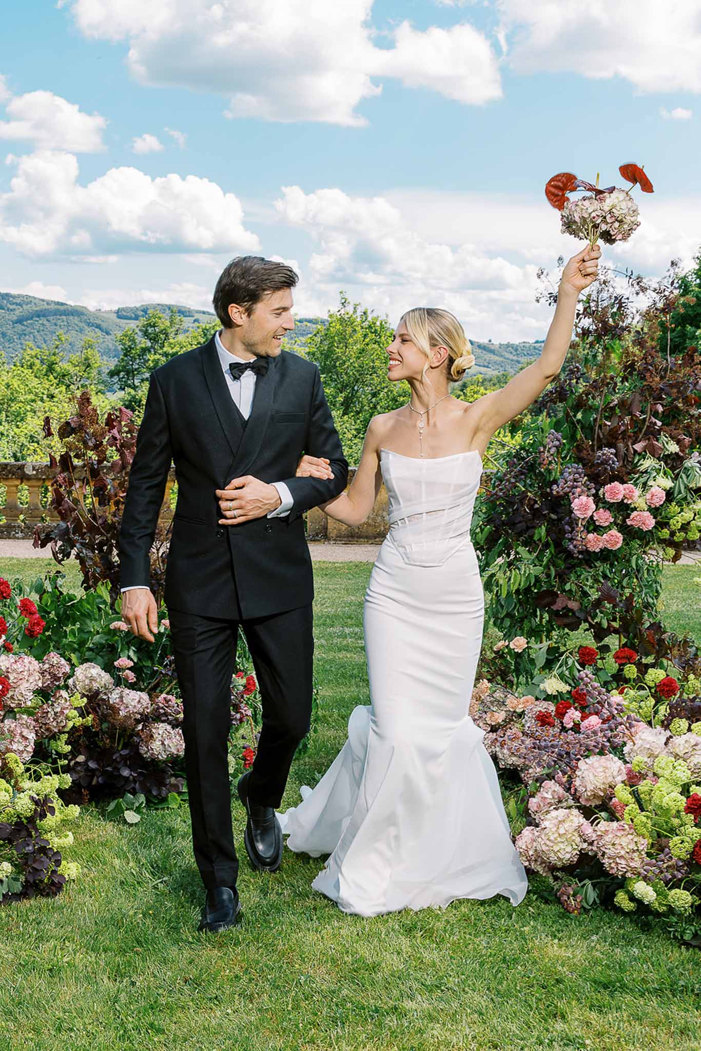 Bride raising blush bouquet with red anthurium overhead beside groom with blush hydrangea and burgundy ground florals